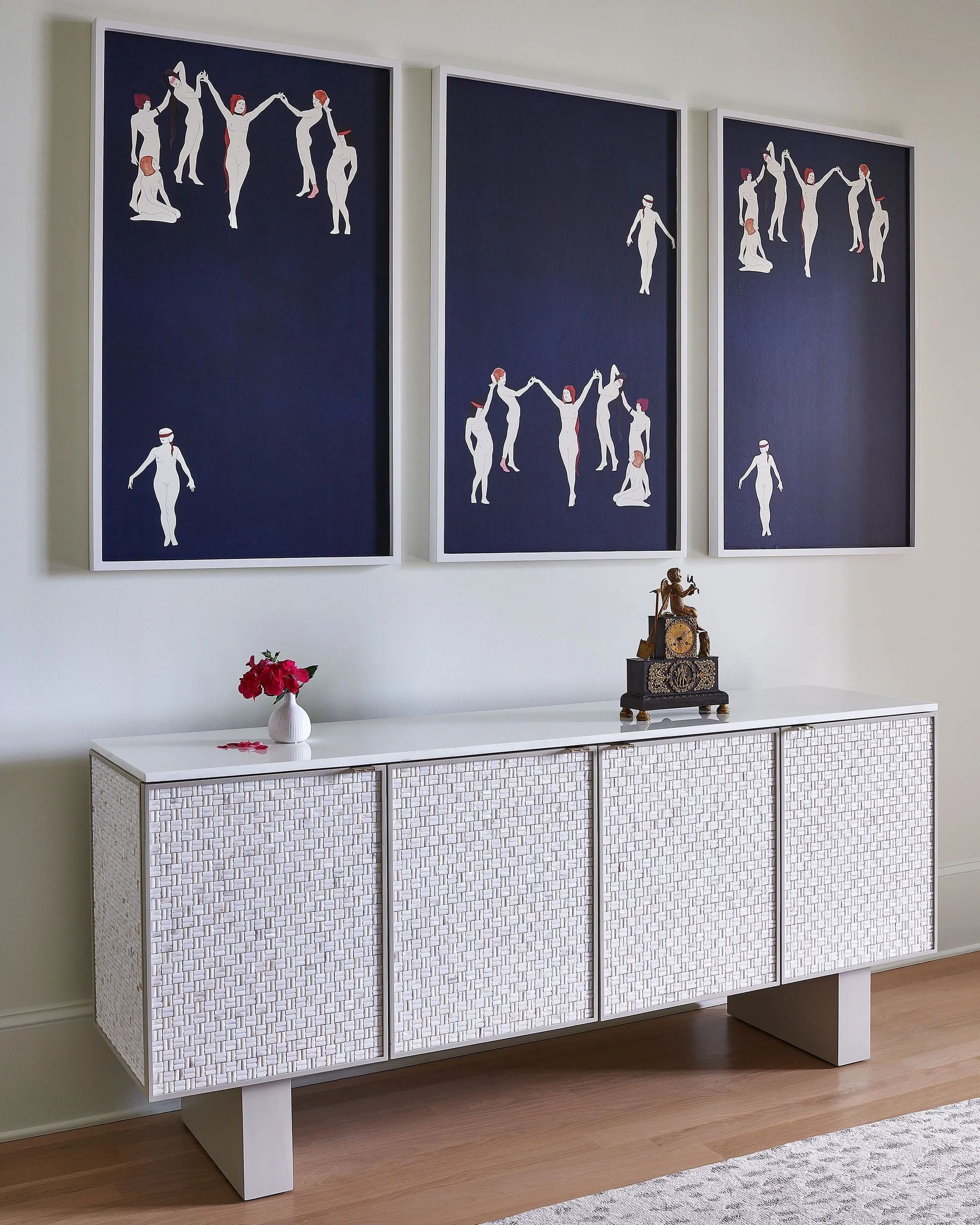 A white four-door console table with woven texture doors sits beneath three large framed navy blue paintings featuring minimalist white figures.