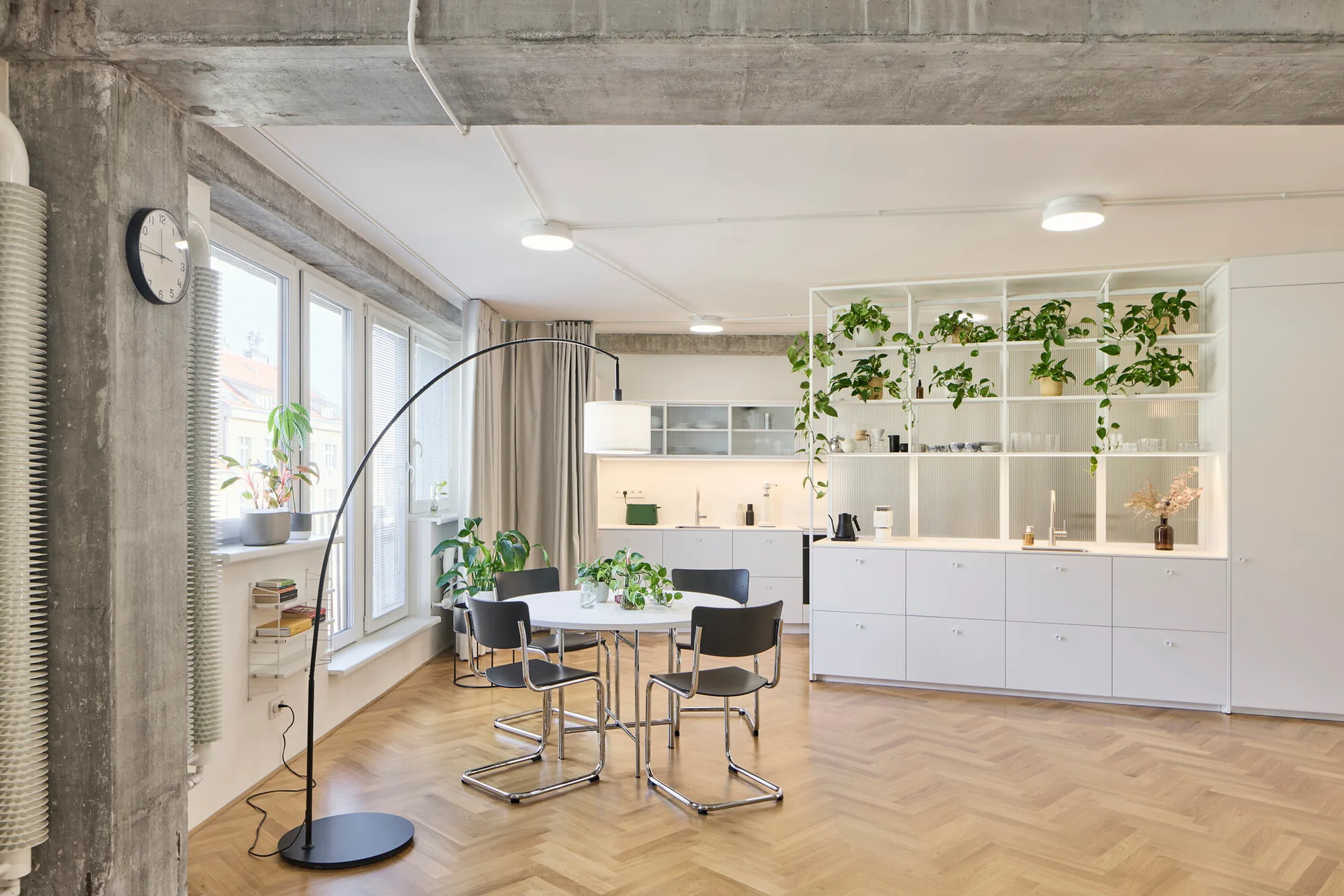 A modern open-plan kitchen and dining area features white cabinetry, an open metal shelving unit with potted plants, a round table, and concrete beams.