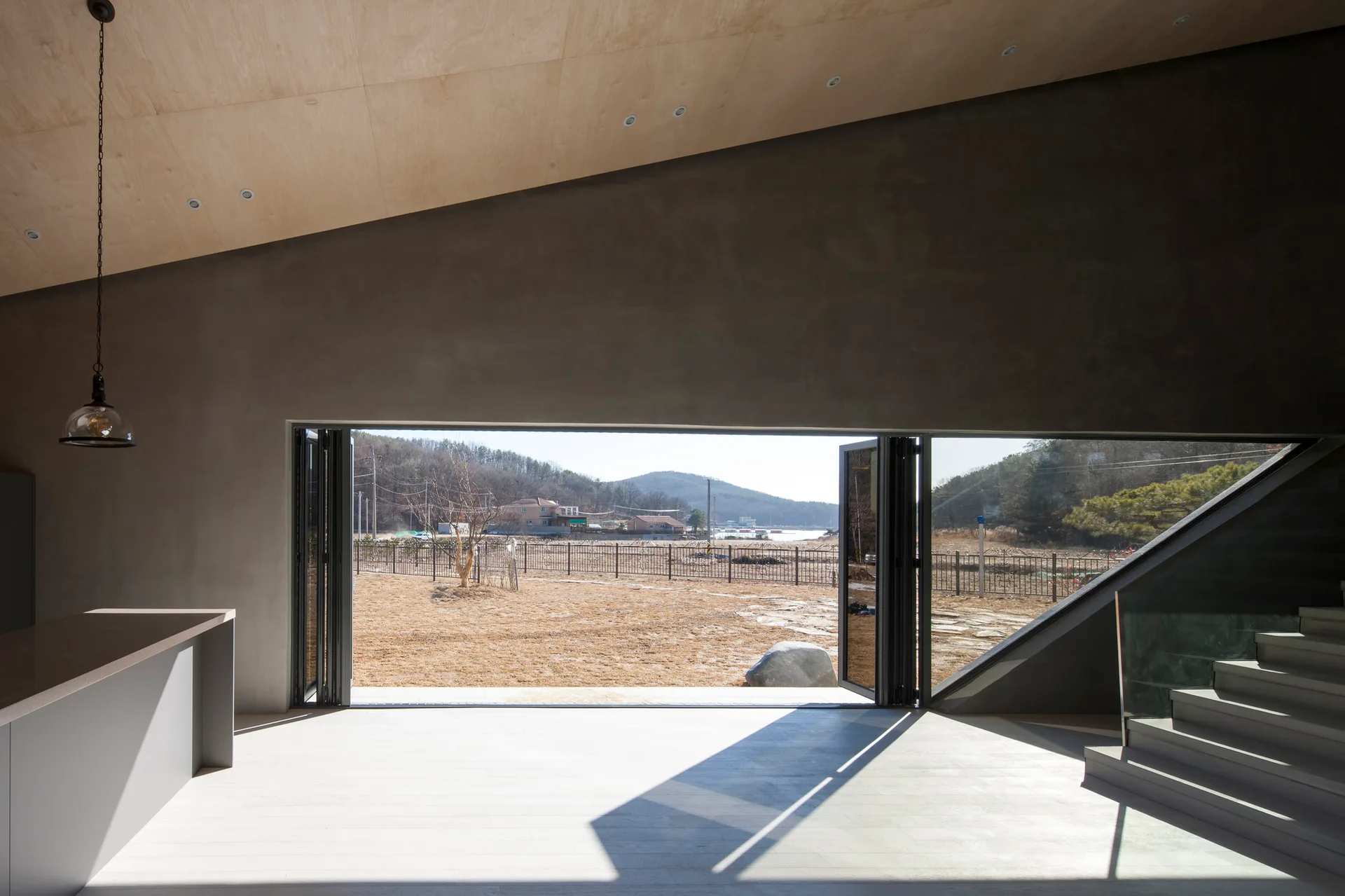 A minimalist open-plan room with a sloped plywood ceiling, smooth grey concrete walls, and a large folding glass door leading outside.