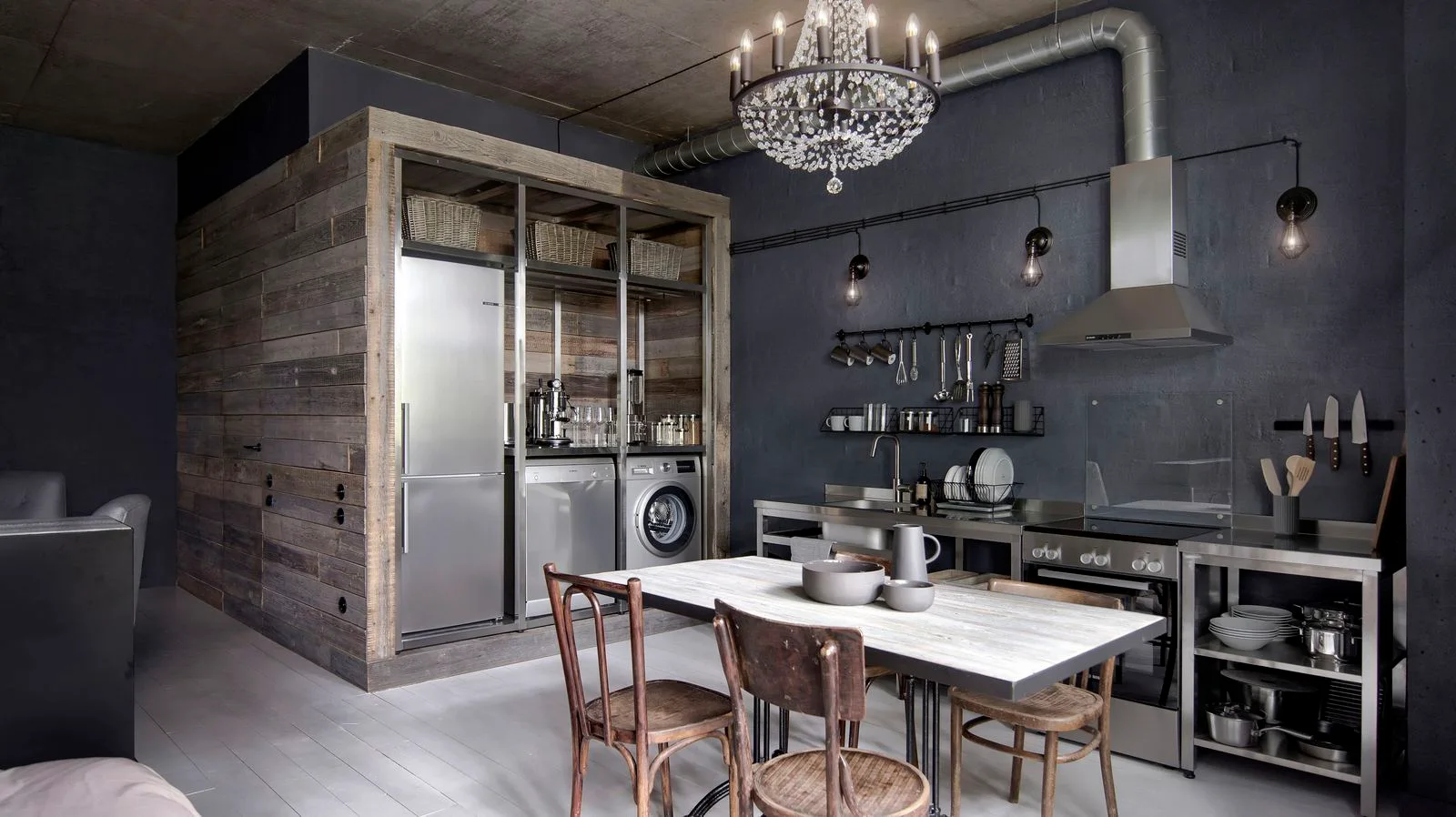 An industrial kitchen featuring dark grey walls, stainless steel counters, a reclaimed wood appliance enclosure, and an ornate crystal chandelier.