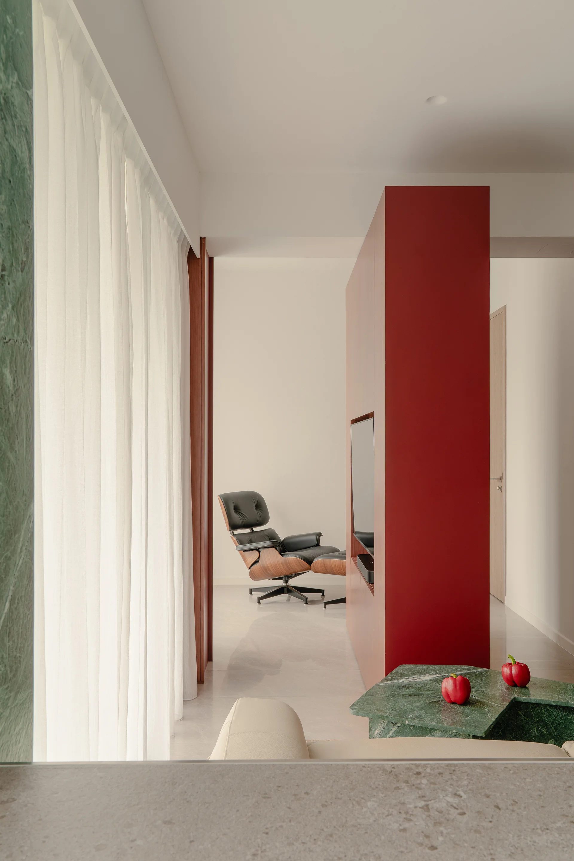 A modern living space features a bold red partition wall with an integrated television, a green marble coffee table, and a black leather lounge chair.