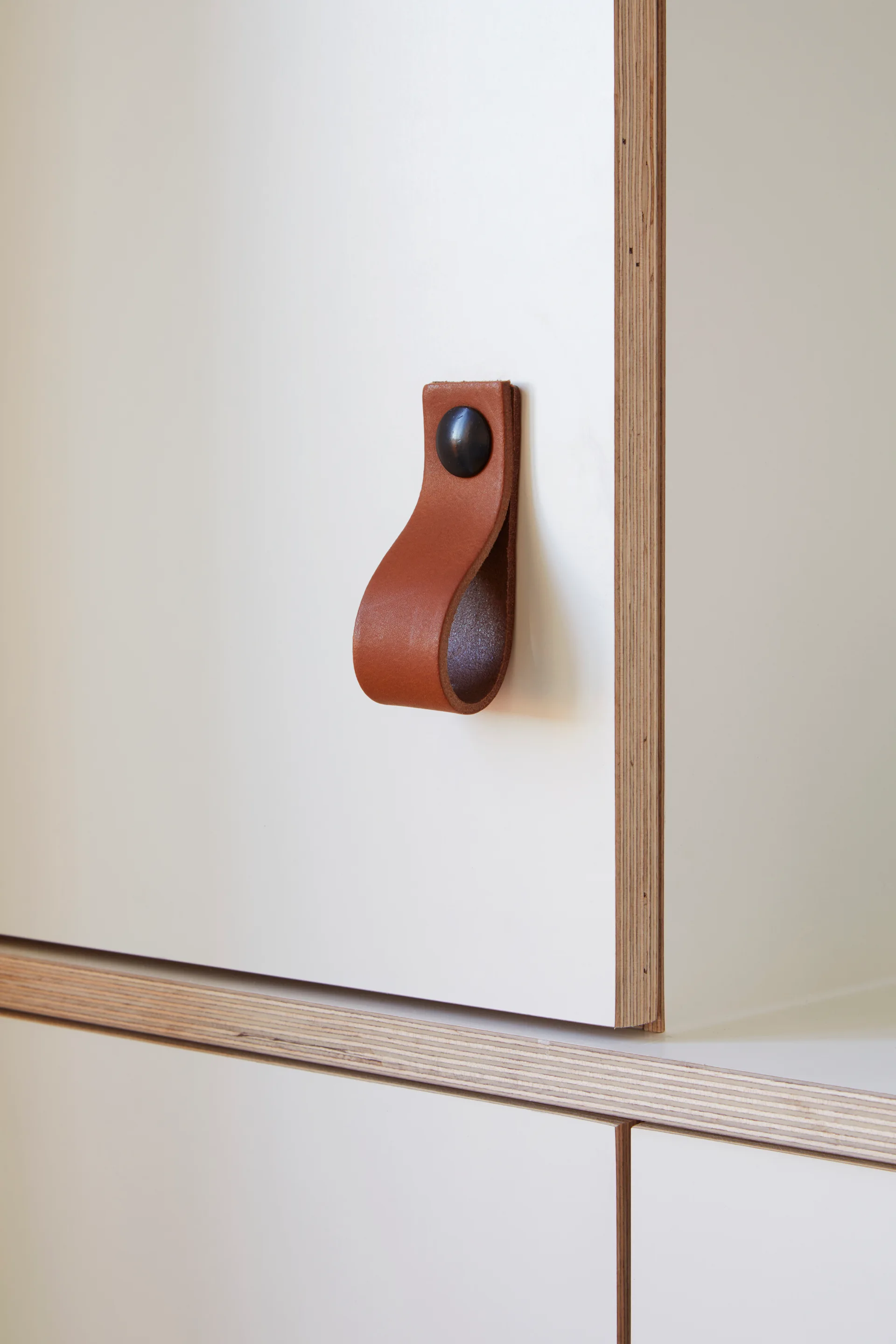 A close-up of a white laminate cabinet door featuring an exposed plywood edge and a brown leather loop strap handle with a dark metal fastener.