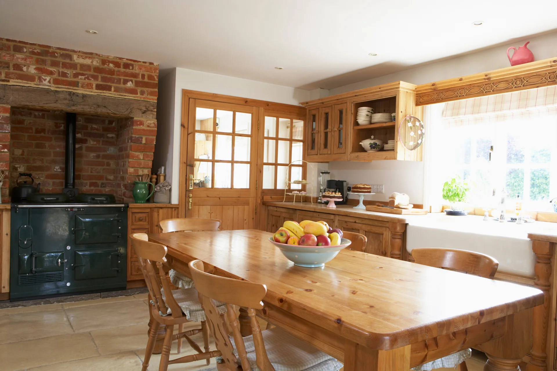 A rustic farmhouse kitchen features a pine dining table and chairs, a dark green range cooker within an exposed brick alcove, and natural wood cabinetry.