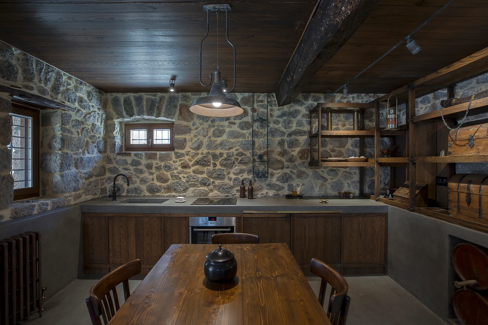A rustic kitchen features exposed stone walls, dark wood ceiling planks with a heavy beam, a wood dining table, and dark wood base cabinets.