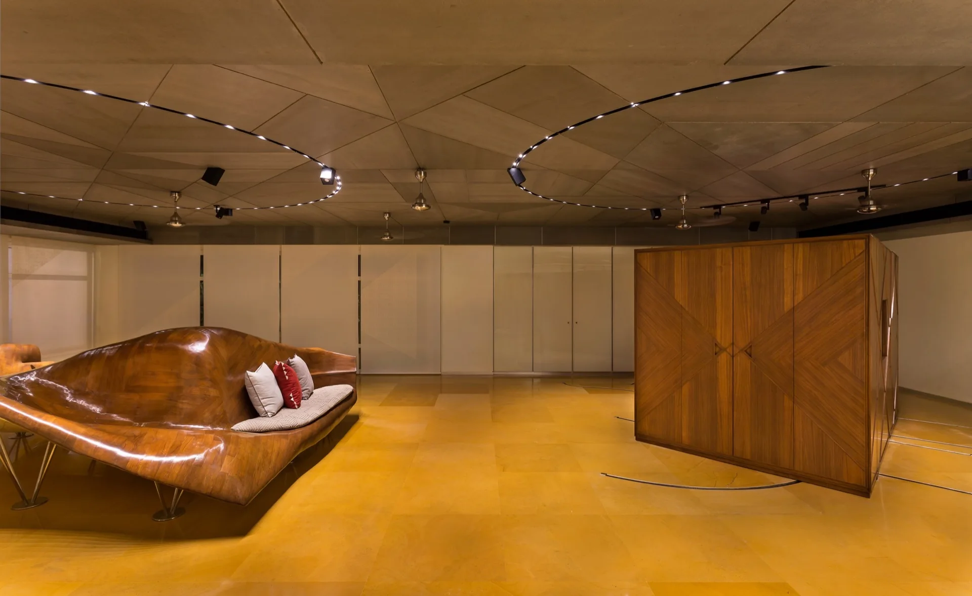A modern minimalist lounge features a glossy curved wooden sofa and a large geometric wooden cabinet beneath circular track lighting on a paneled ceiling.