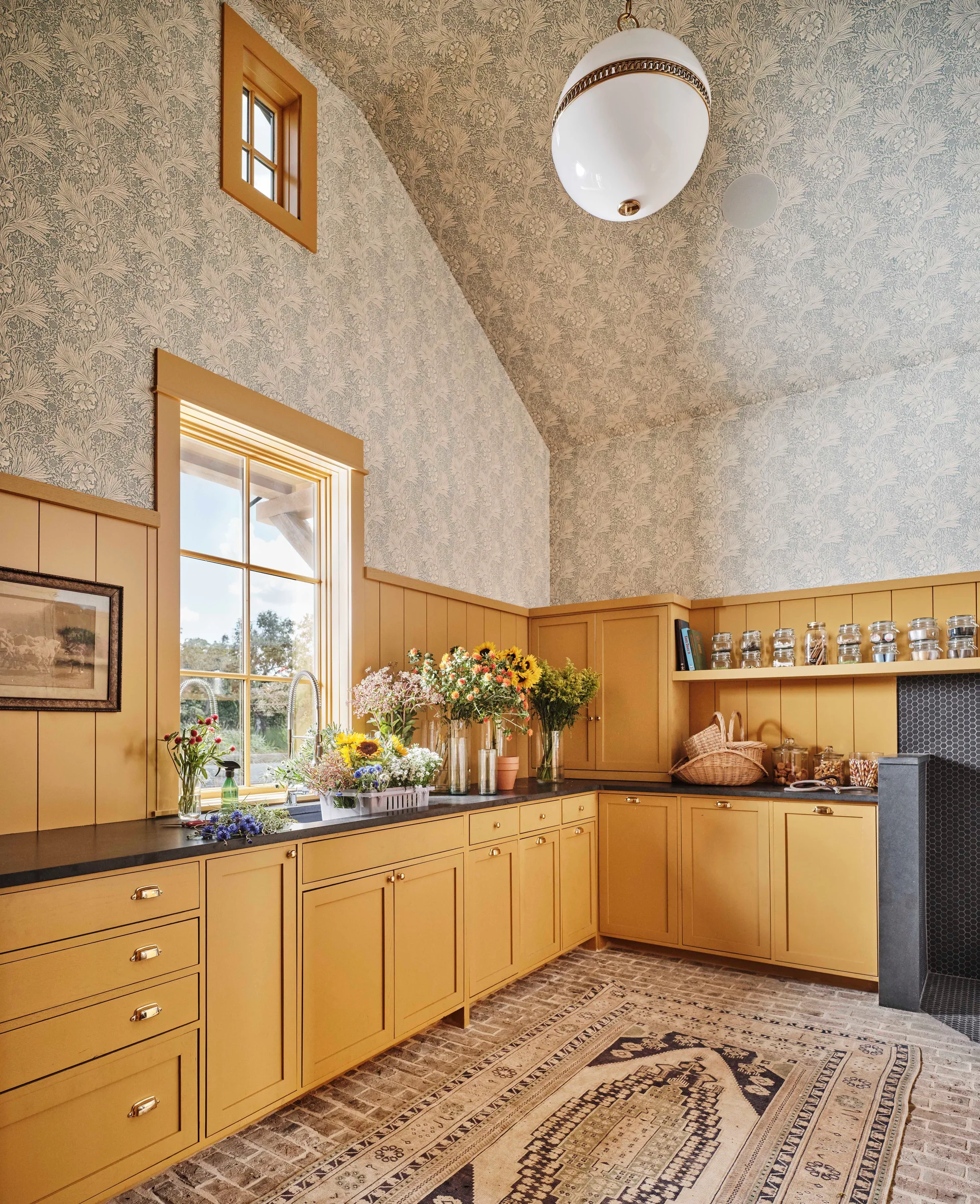 A utility room features mustard yellow cabinets, vertical wood paneling, a brick floor, and floral patterned wallpaper on the walls and vaulted ceiling.