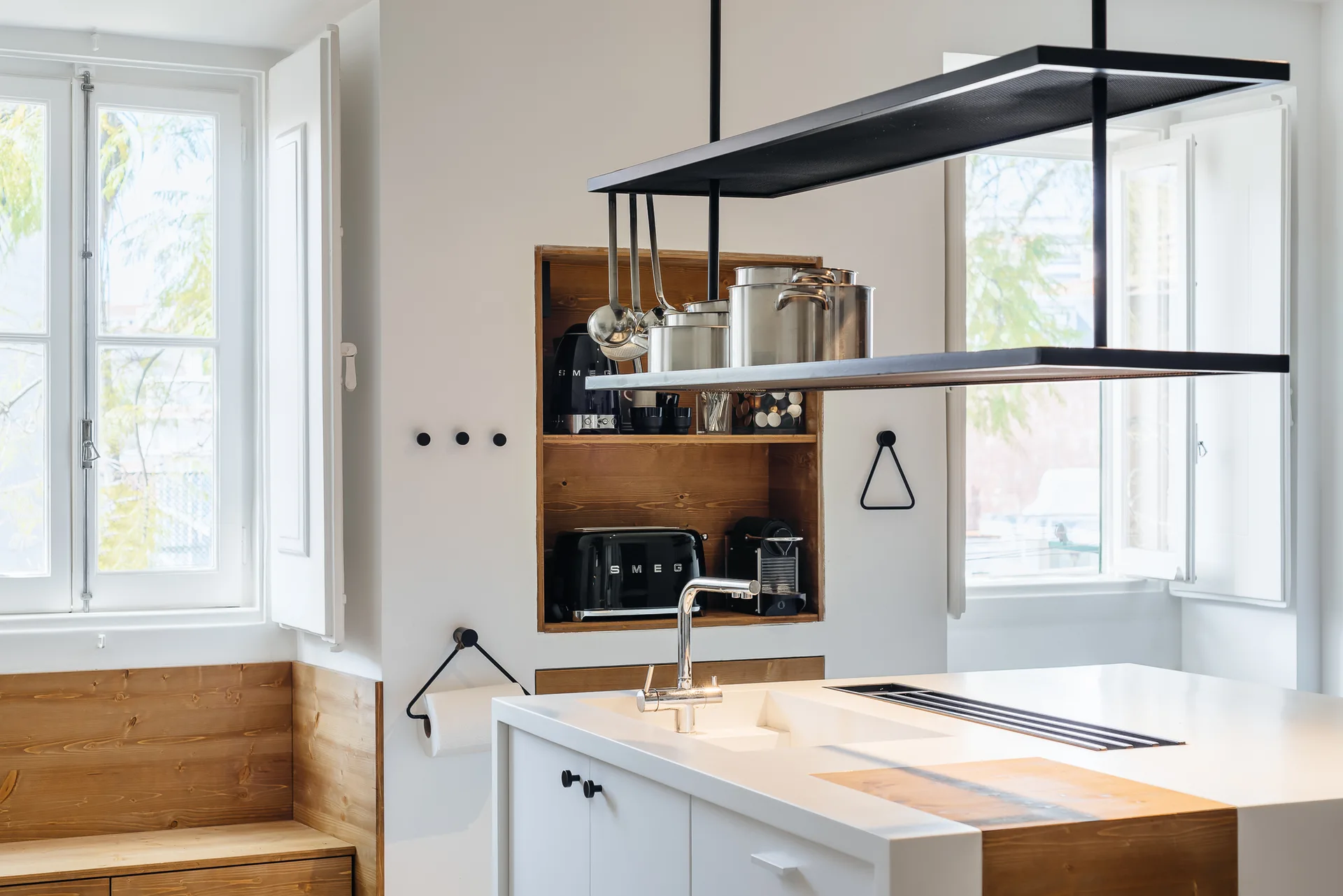A modern white kitchen island with a chrome faucet stands in front of a recessed wood-lined wall niche filled with black appliances.