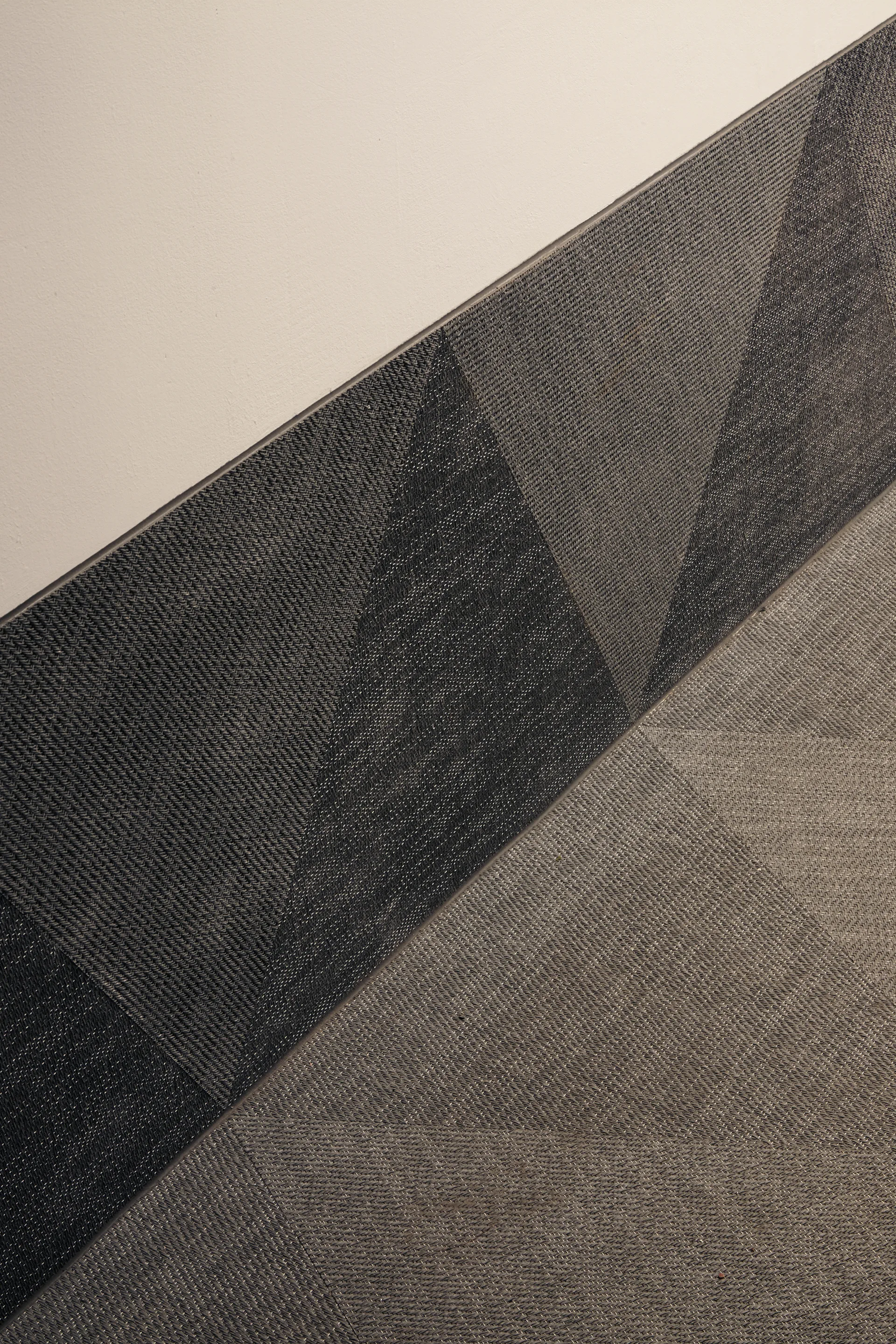 A close-up of a charcoal grey woven fabric wall panel featuring a geometric triangle pattern meeting a smooth off-white wall along a diagonal line.