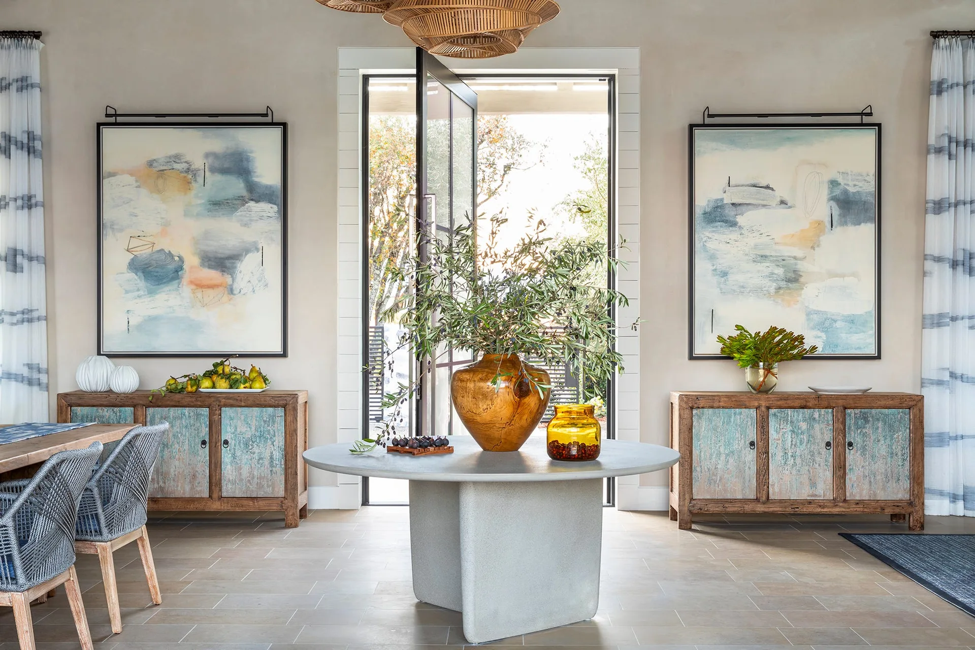 A bright entryway features a round stone table flanked by two matching distressed wood credenzas topped with large framed abstract paintings.