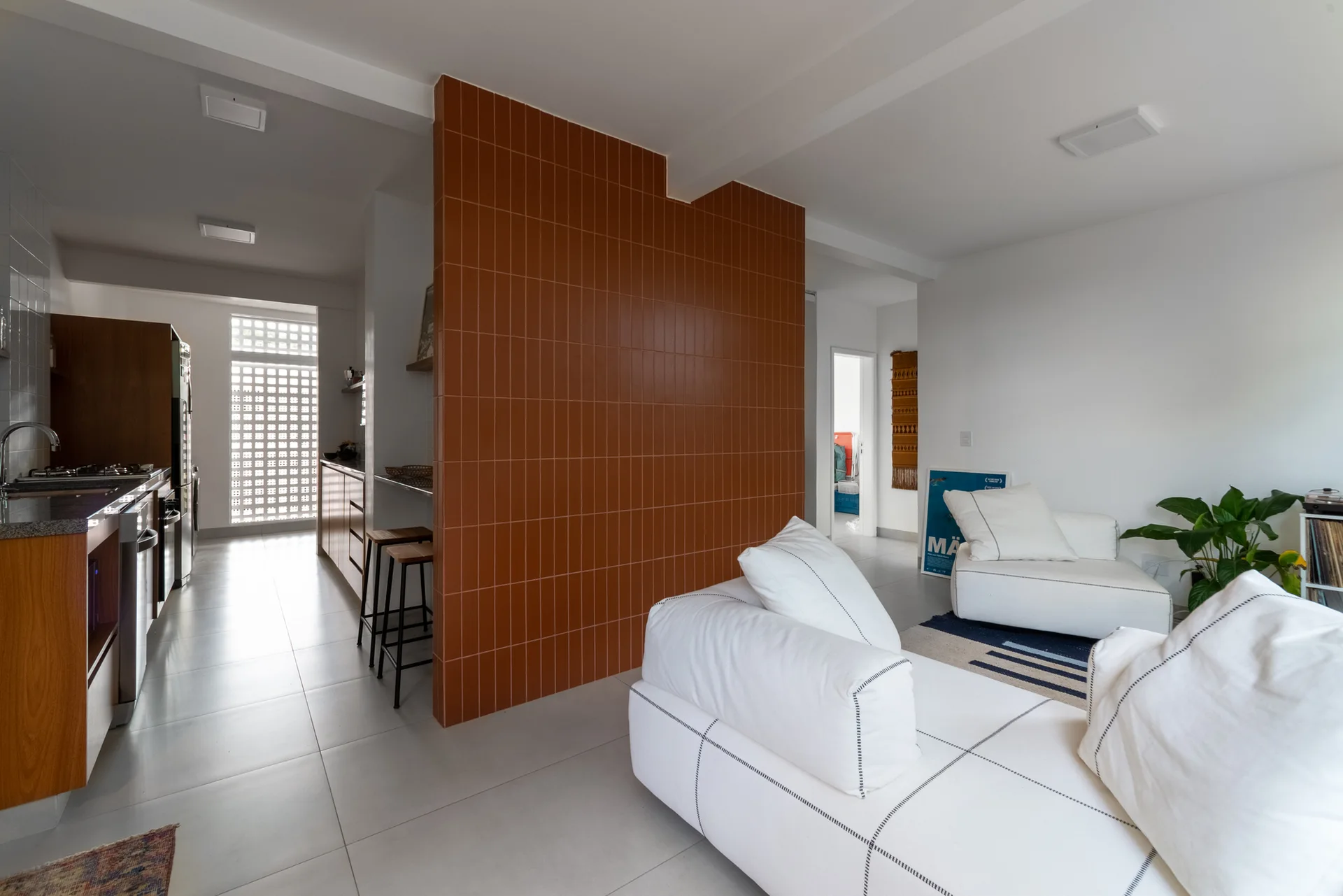 An open-plan living area features a white sectional sofa, a tall partition wall covered in terracotta ceramic tiles, and a kitchen with wood cabinets.