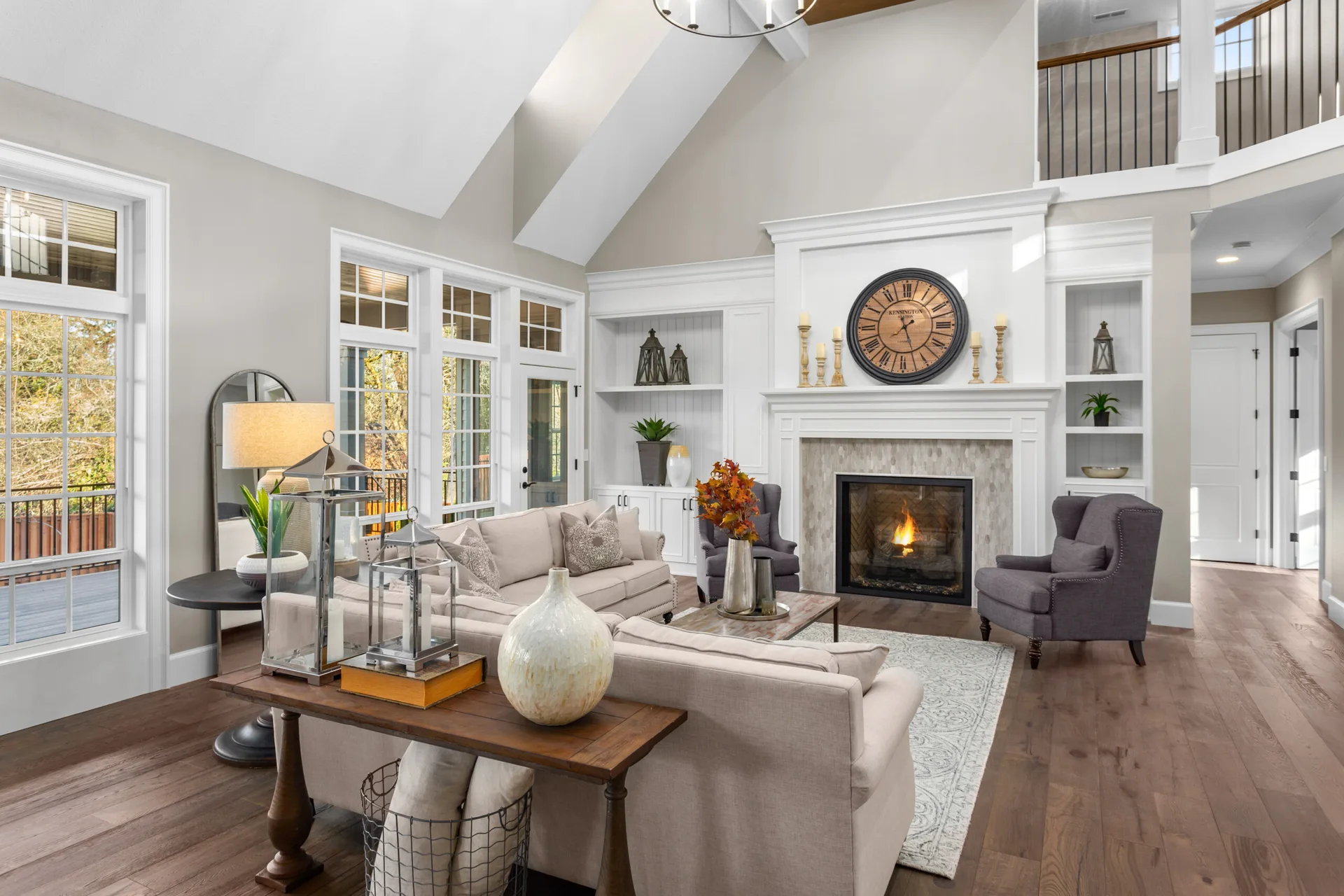 A spacious living room features a fireplace flanked by white built-in shelves, a beige sofa, and a large round wall clock under a vaulted ceiling.