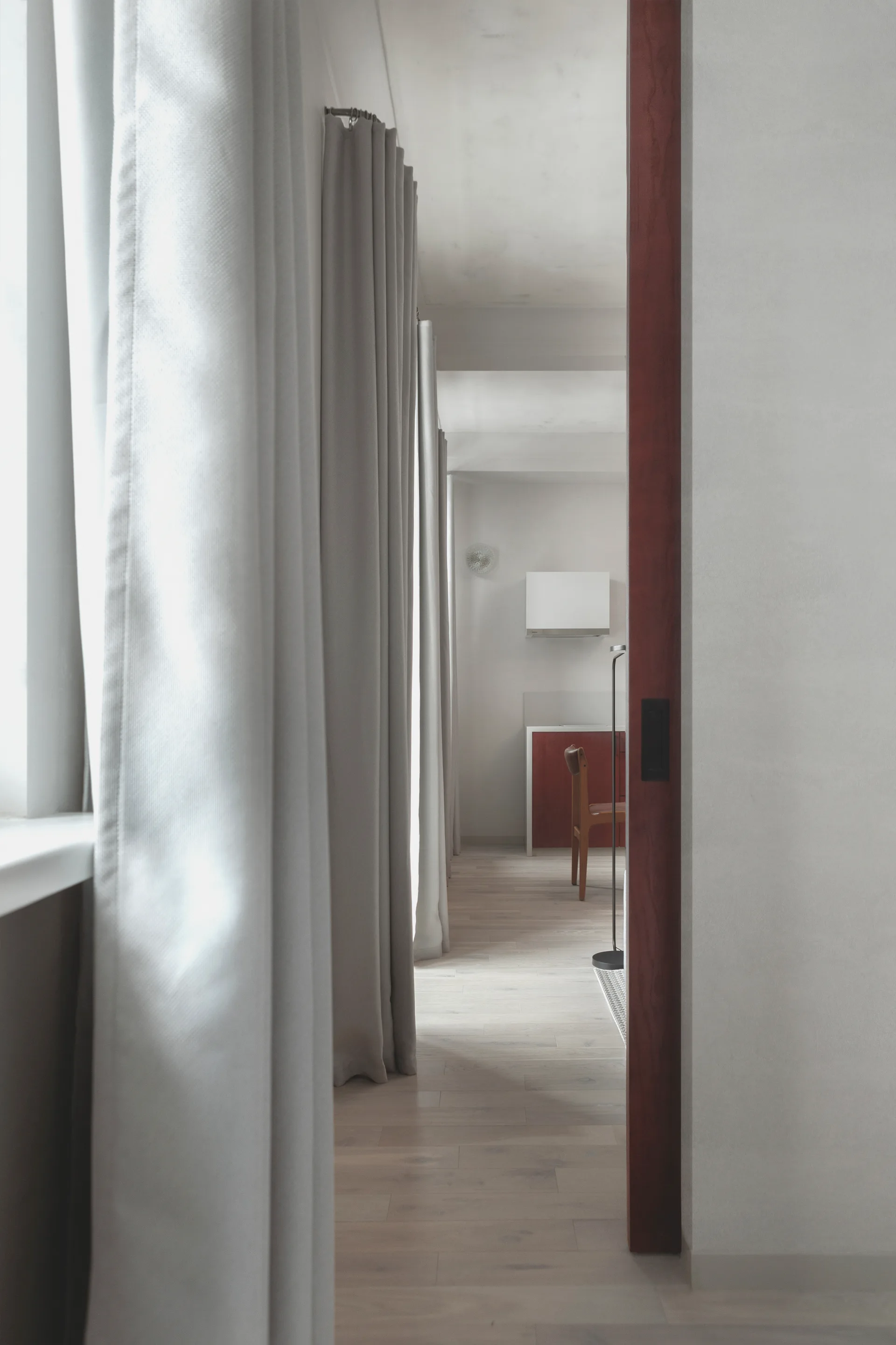 A view through a dark wood-framed doorway down a hallway with long grey curtains on light wood flooring leading to a kitchen in the distance.