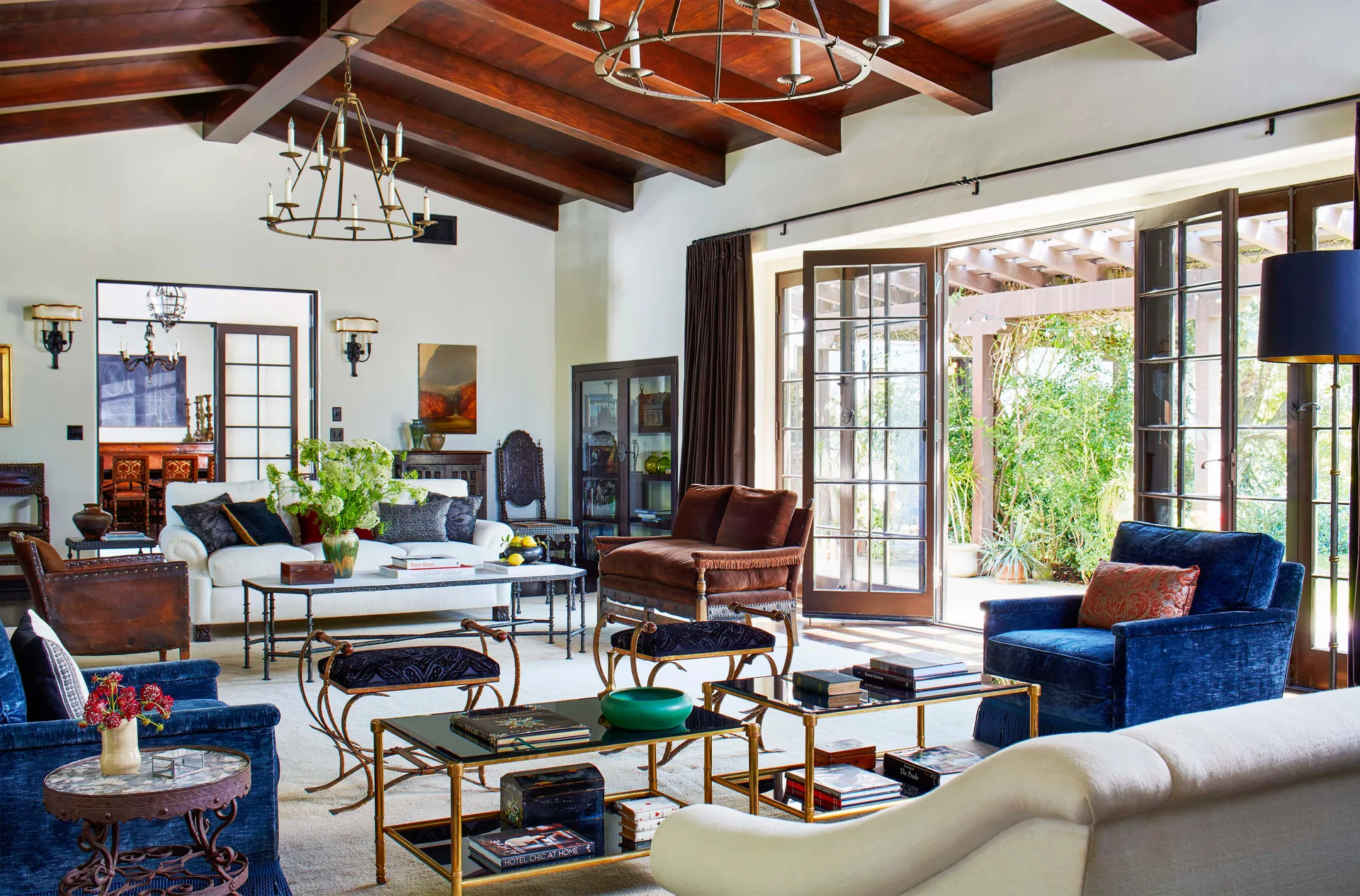 A spacious living room features exposed dark wood ceiling beams, cream walls, blue velvet armchairs, and glass doors opening to a lush garden patio.