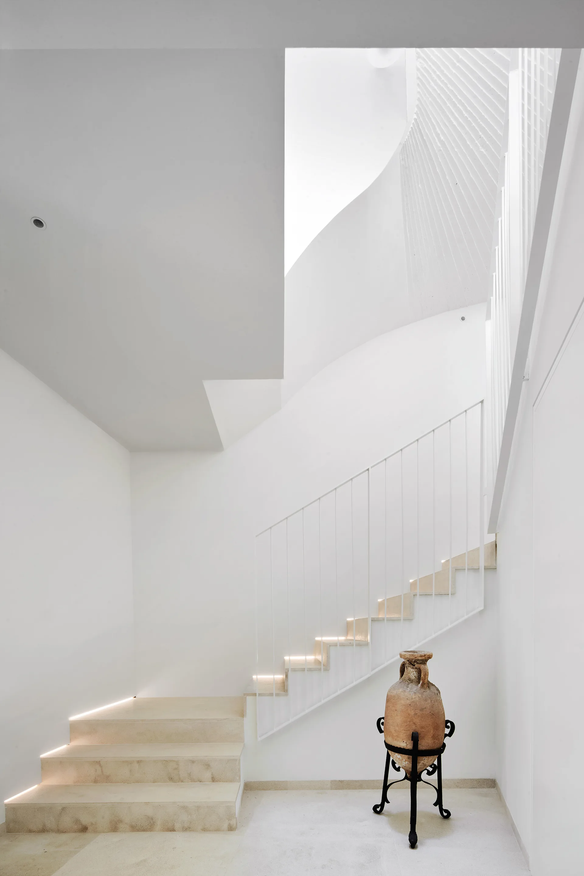 A minimalist white staircase features stone-like steps lit by integrated LED strips and a large antique clay urn on a black stand.