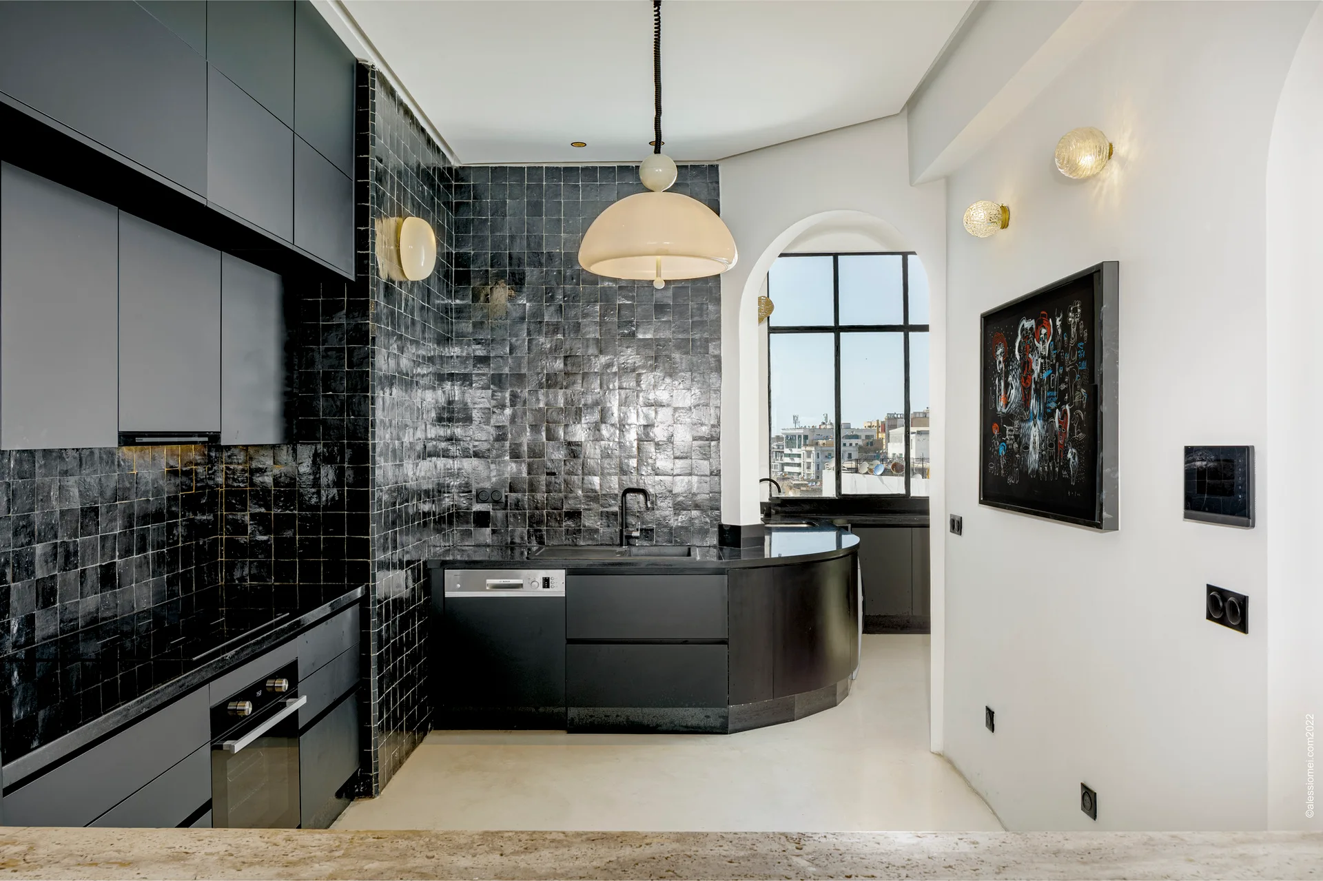 A modern kitchen features matte black cabinetry, a matching curved counter, glossy square tiled walls, and an arched doorway leading to a grid-paneled window.