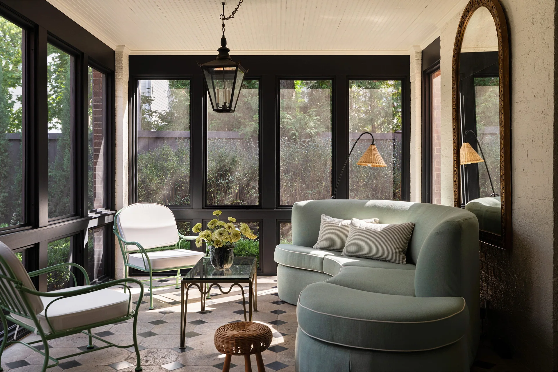 A sunlit screened porch features a curved light blue sofa, green metal armchairs, a large arched wall mirror, and black-trimmed windows.