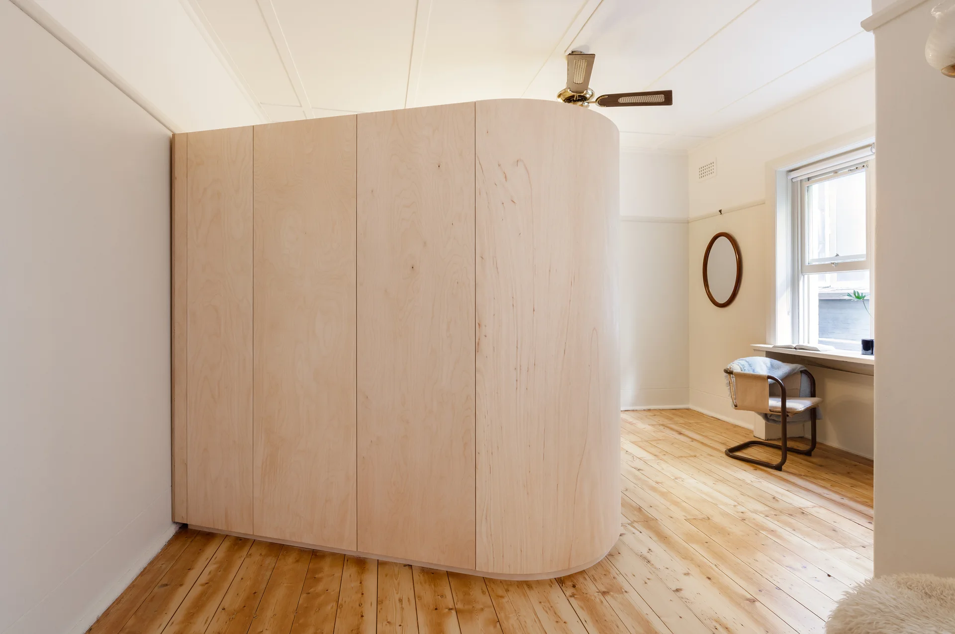A large curved plywood partition wall stands on light hardwood floors in a white minimalist room with a window and a ceiling fan.