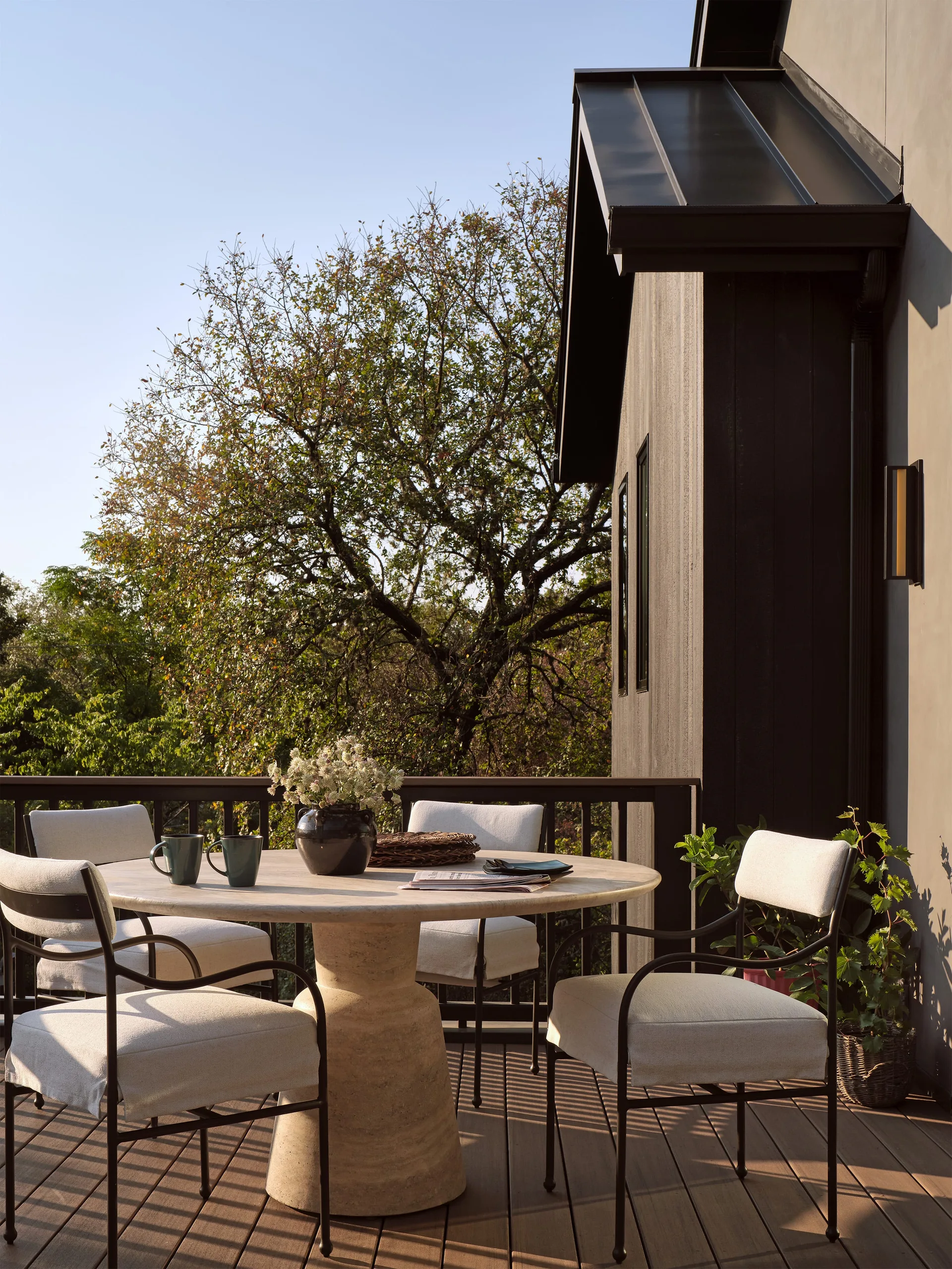 A modern outdoor dining set featuring a round stone pedestal table and four beige upholstered chairs on a dark wood deck next to a dark house wall.