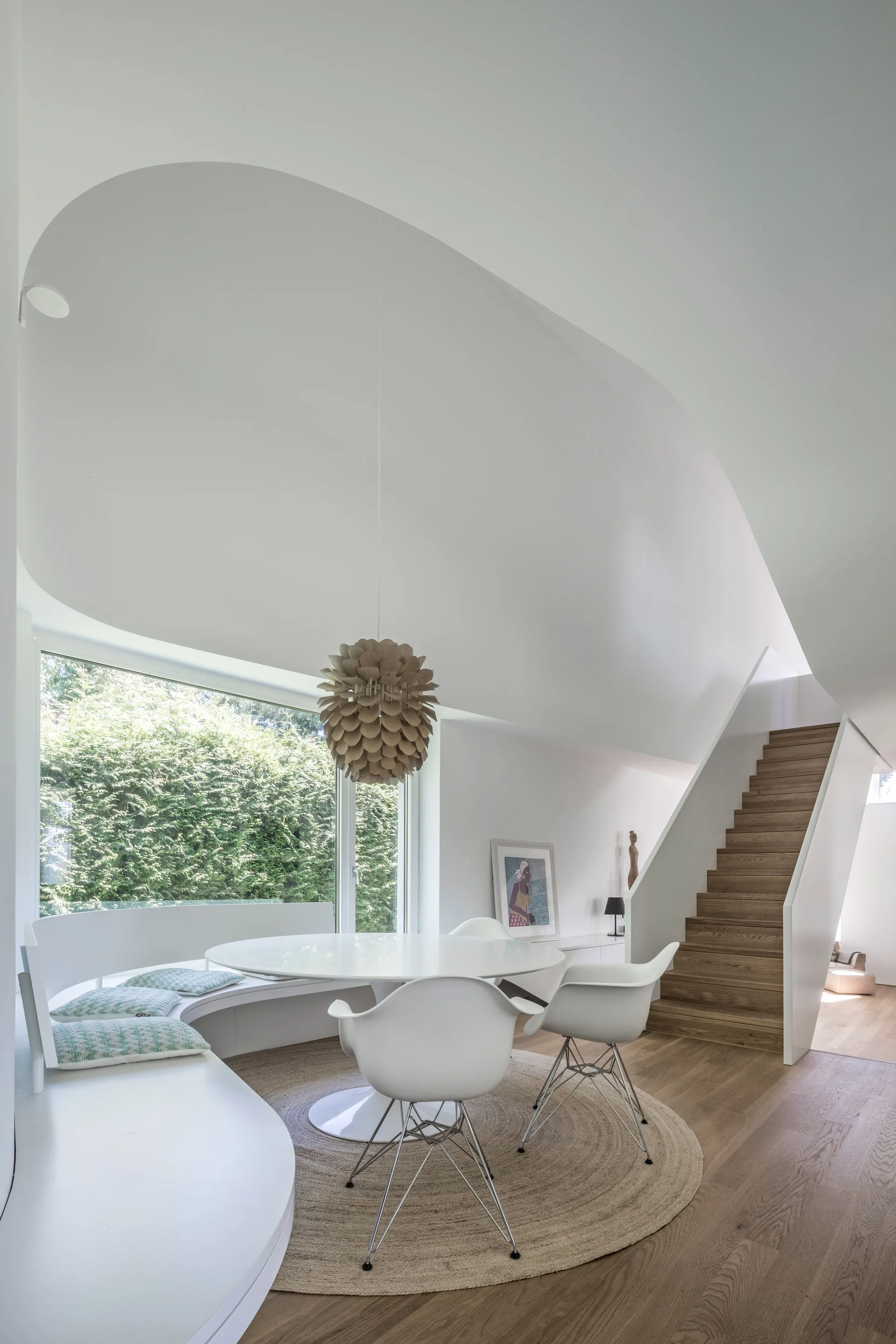 A bright dining room features a curved white built-in banquette, a circular dining table with white chairs, and an adjacent wood-tread staircase.