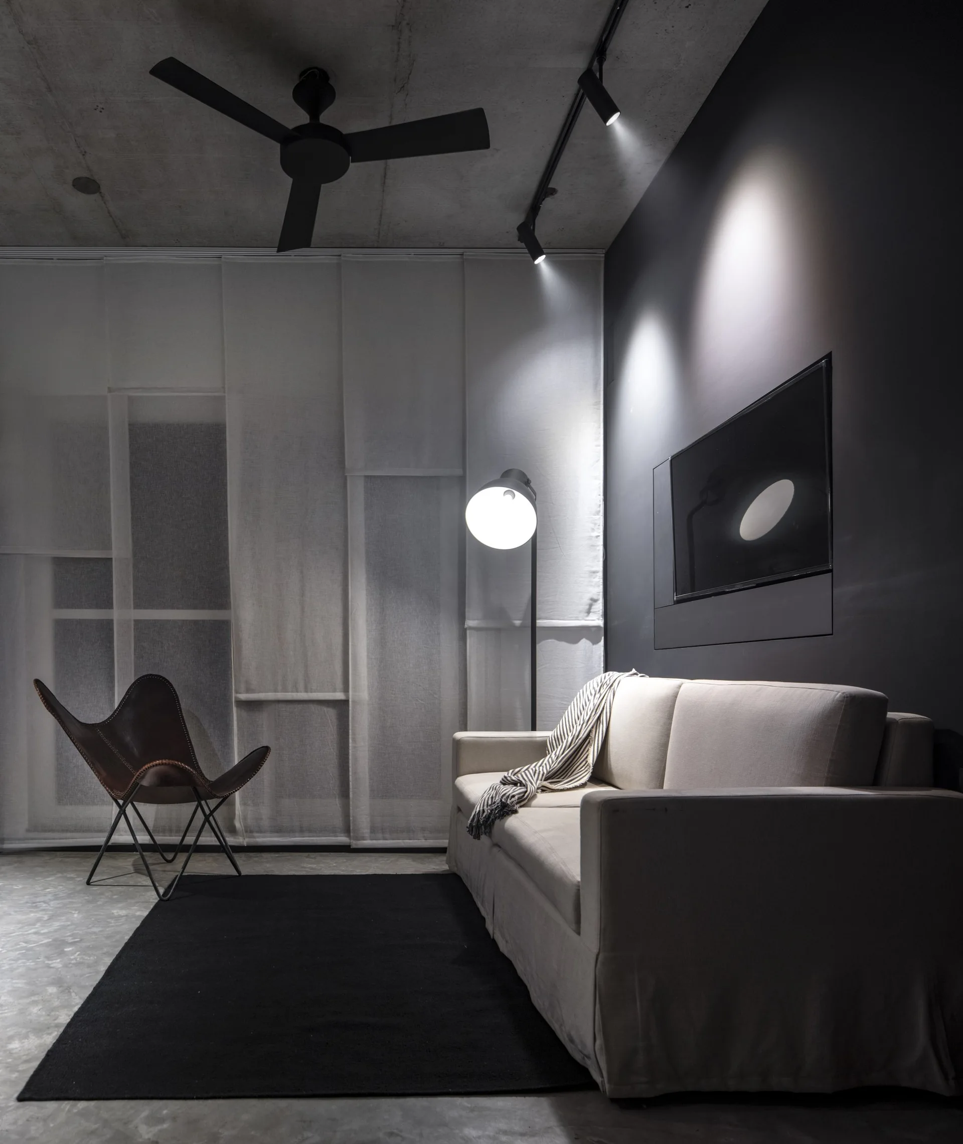 A minimalist living room featuring a cream sofa, a brown leather butterfly chair, a dark accent wall, and a raw concrete ceiling.
