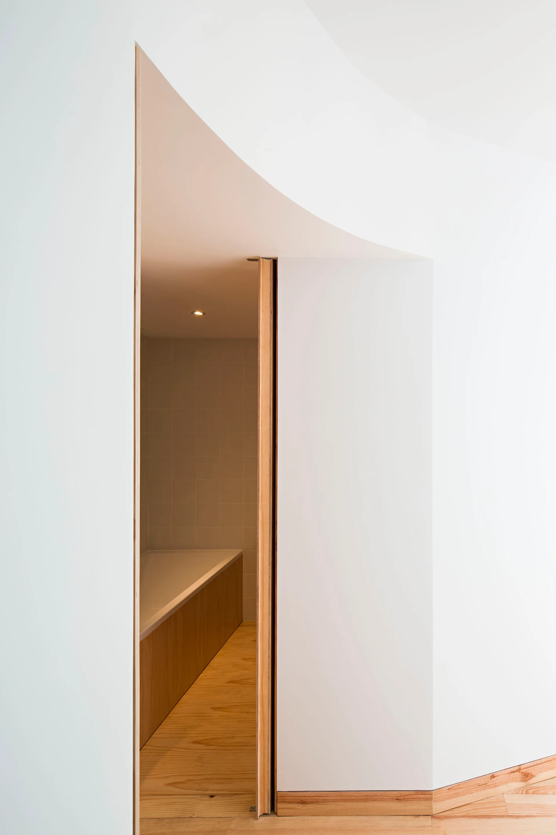 A minimalist white entryway with a curved wall and light wood trim opens to a bathroom featuring a wood-paneled bathtub.