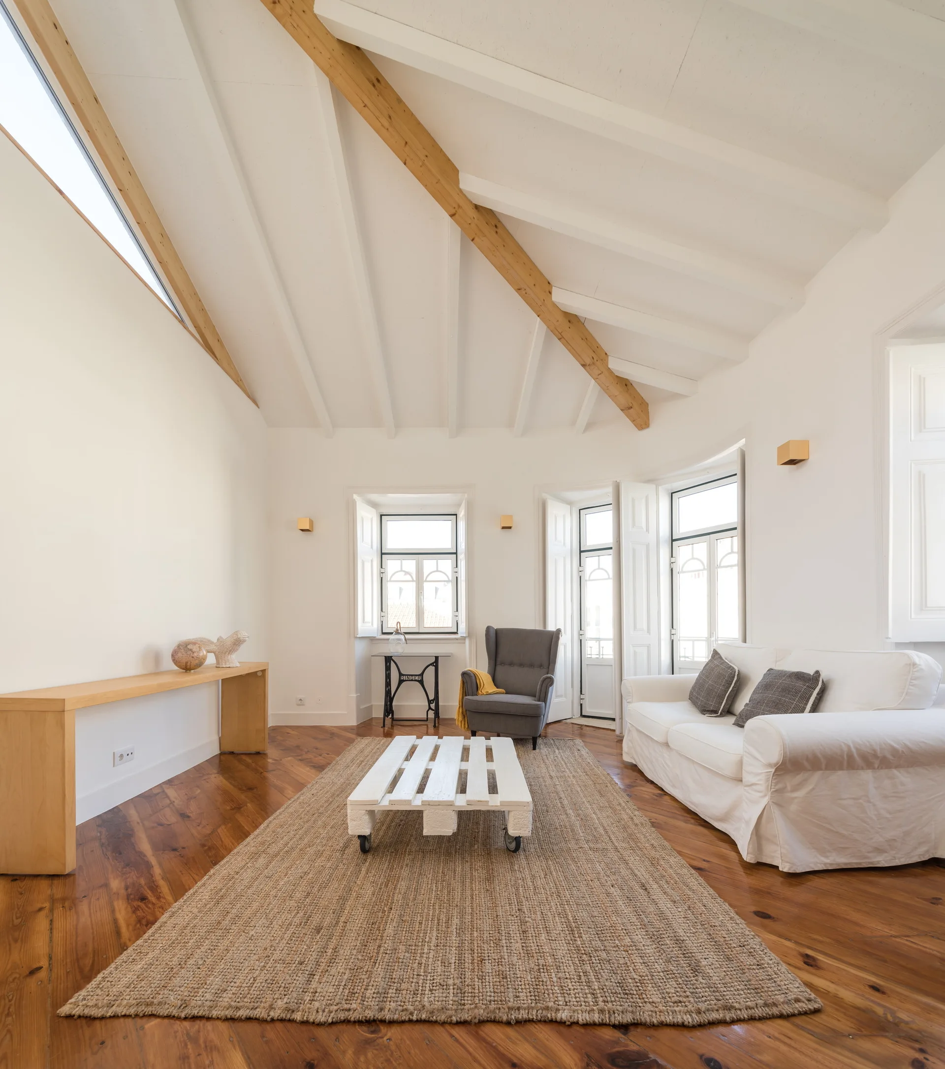 A bright living room with a vaulted ceiling, exposed wood beams, a white sofa, and a woven jute rug.