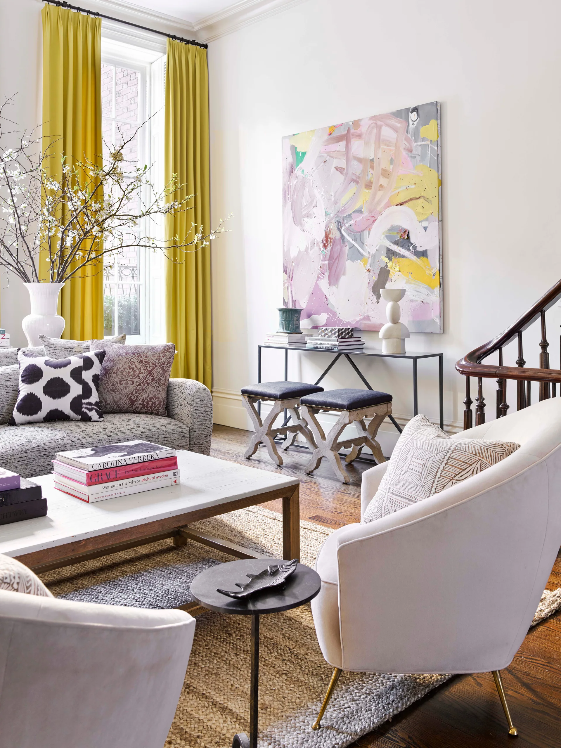 An eclectic living room featuring a grey sofa, white armchairs on a jute rug, a large abstract painting, and bold yellow curtains.