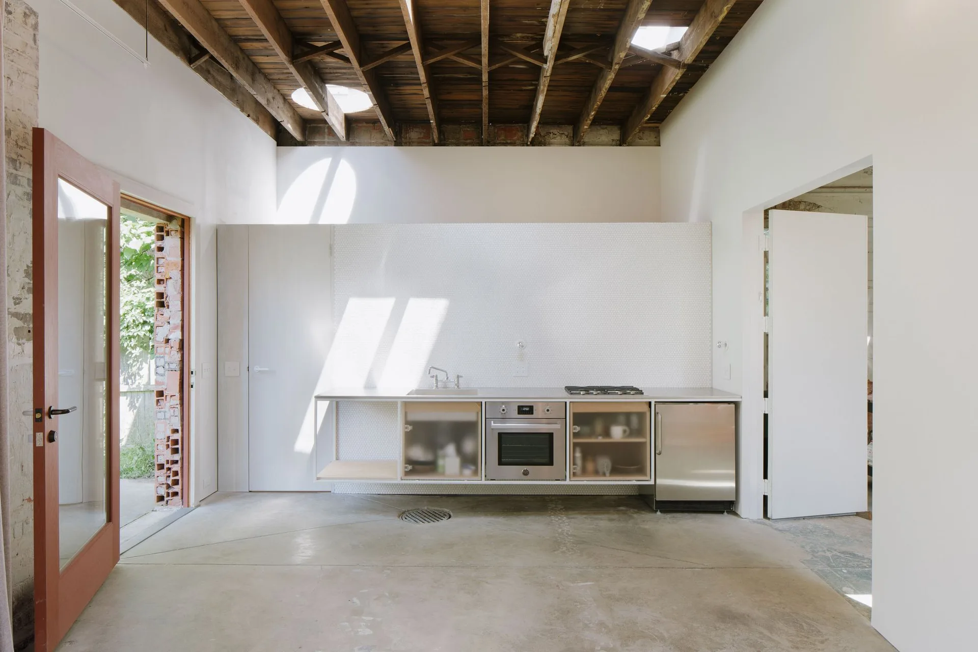A minimalist kitchen features a floating stainless steel counter against a white penny tile wall beneath an exposed raw timber ceiling with skylights.