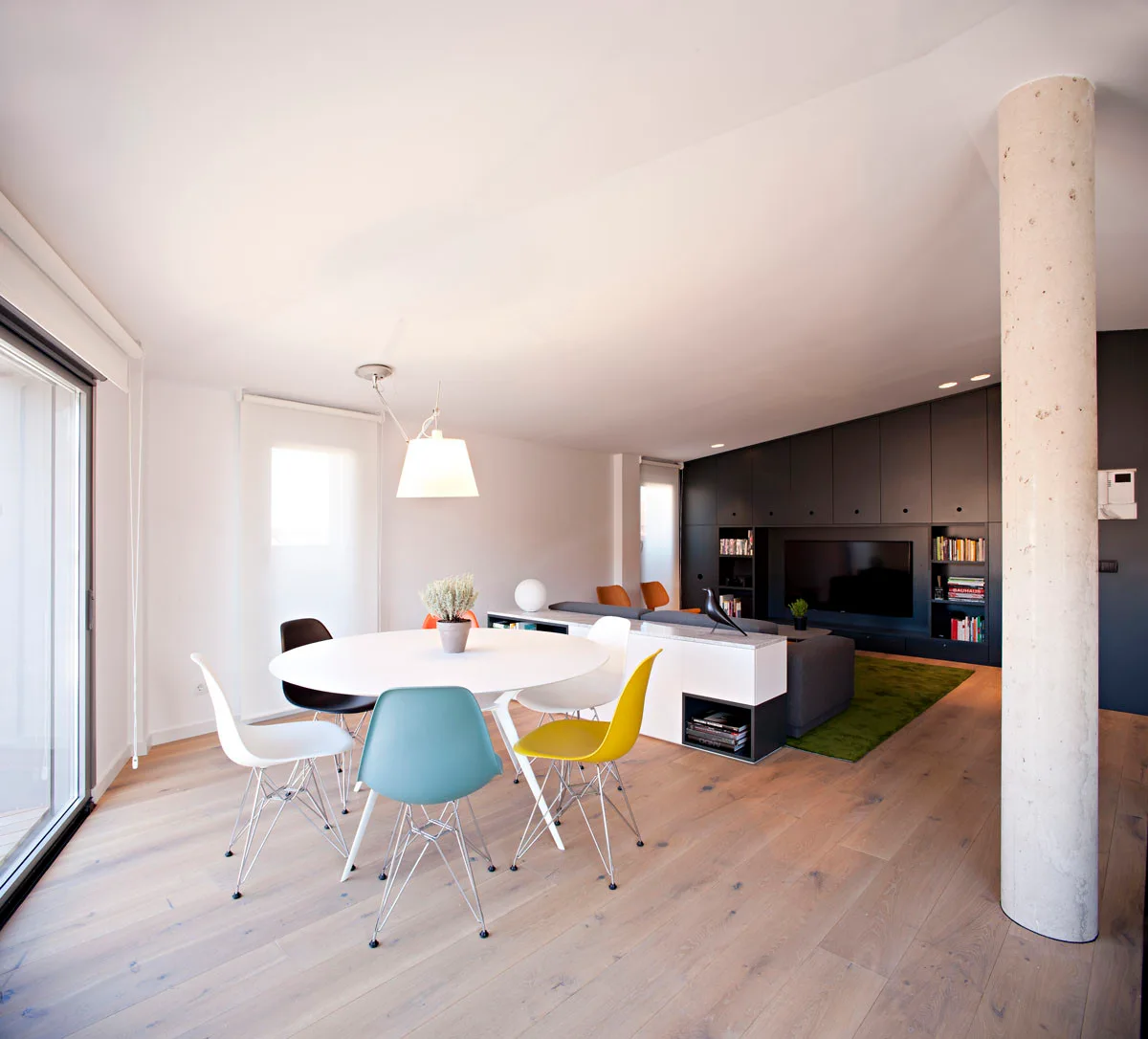 A modern open-plan dining room features a round white table with colorful mid-century shell chairs, a concrete column, and a dark storage wall.