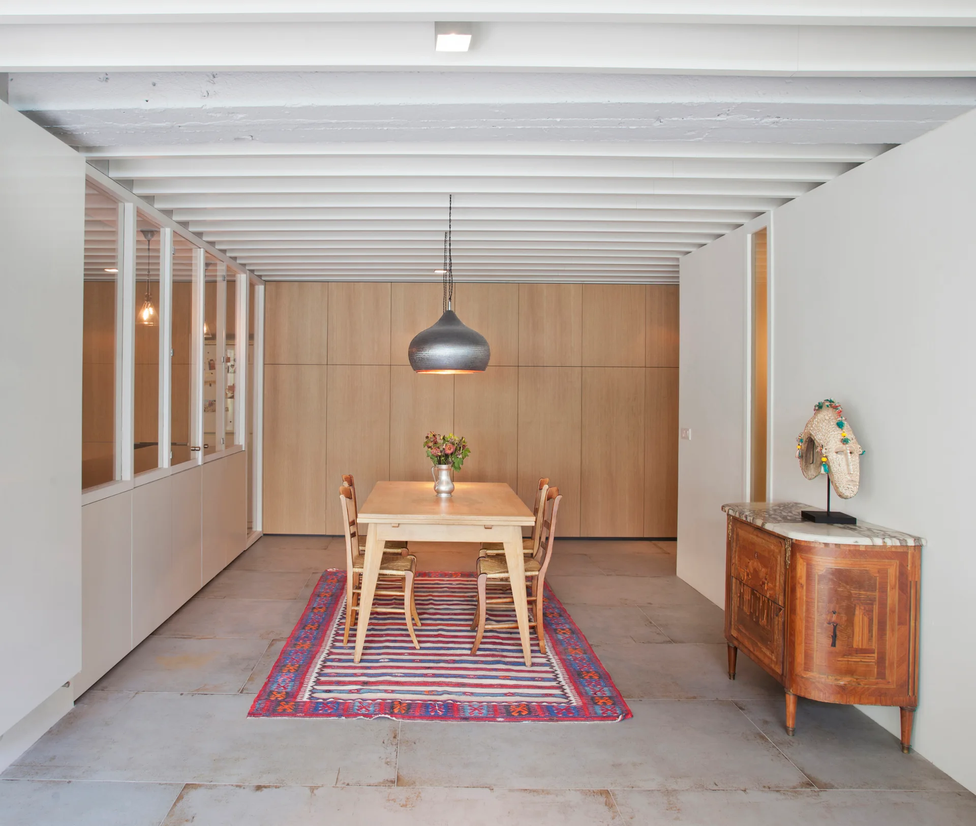 A light wood dining table with chairs sits on a striped red rug beneath white ceiling beams, next to a classic wooden sideboard with marble top.