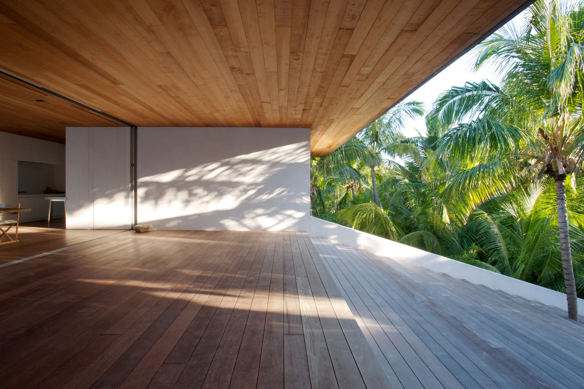 A wooden deck with a matching wood-plank ceiling features a white wall showing palm leaf shadows, adjacent to lush green palm trees.