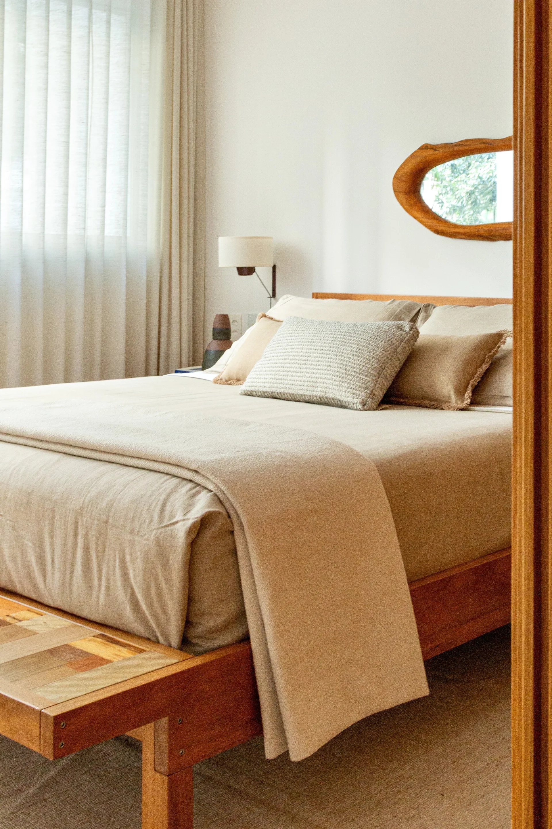 A wooden platform bed with neutral beige linens and pillows is positioned next to a sheer curtained window under an organic wood-framed mirror.