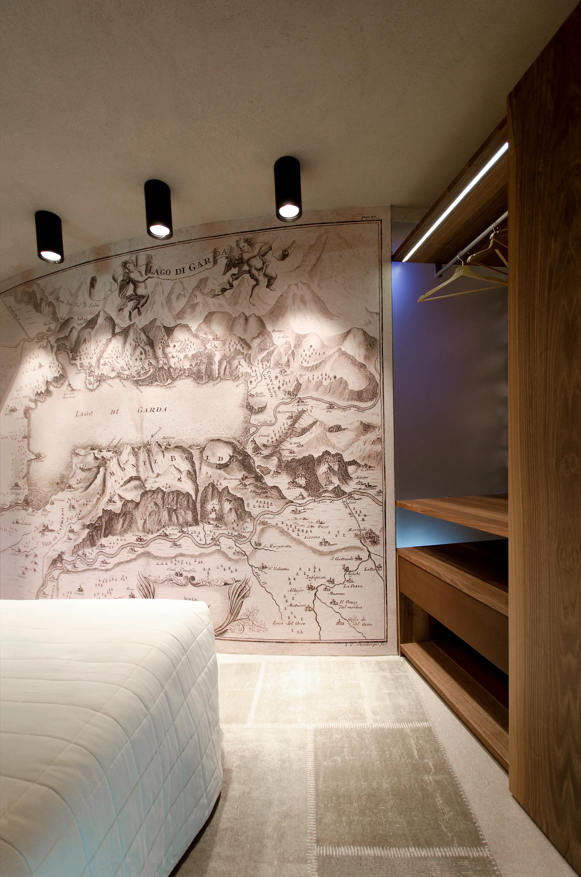 A bedroom features a large vintage map mural on the wall illuminated by ceiling spotlights next to a wooden wardrobe closet with integrated LED lighting.