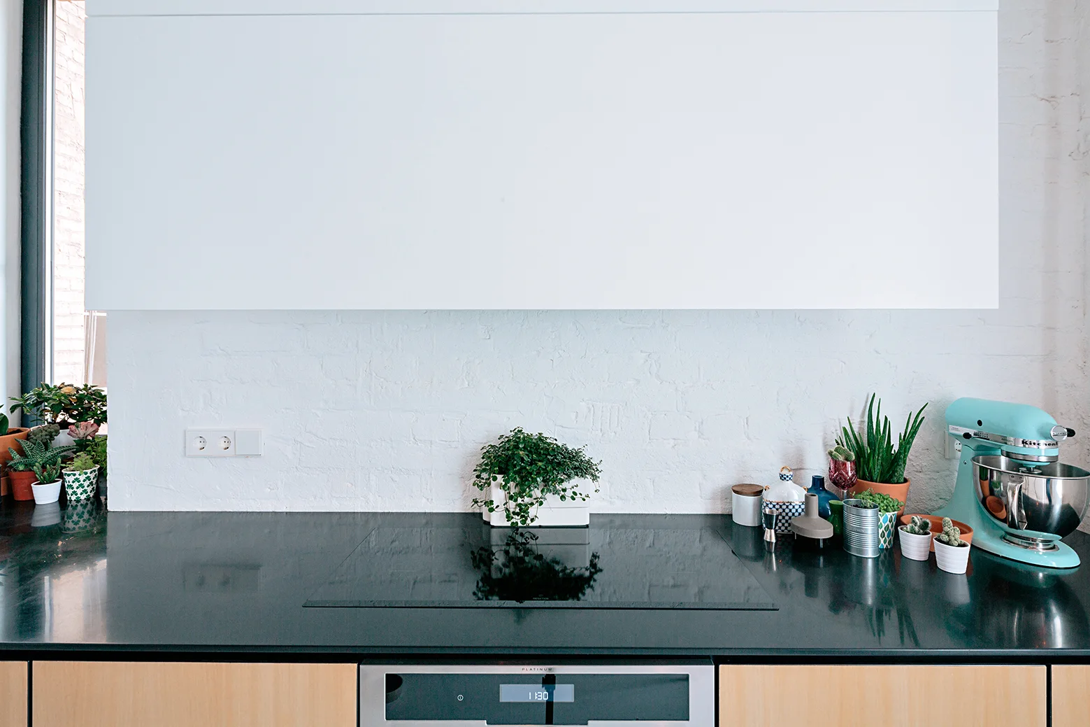 A dark kitchen countertop with an induction cooktop sits below a painted white brick backsplash, flanked by potted plants and a light blue mixer.