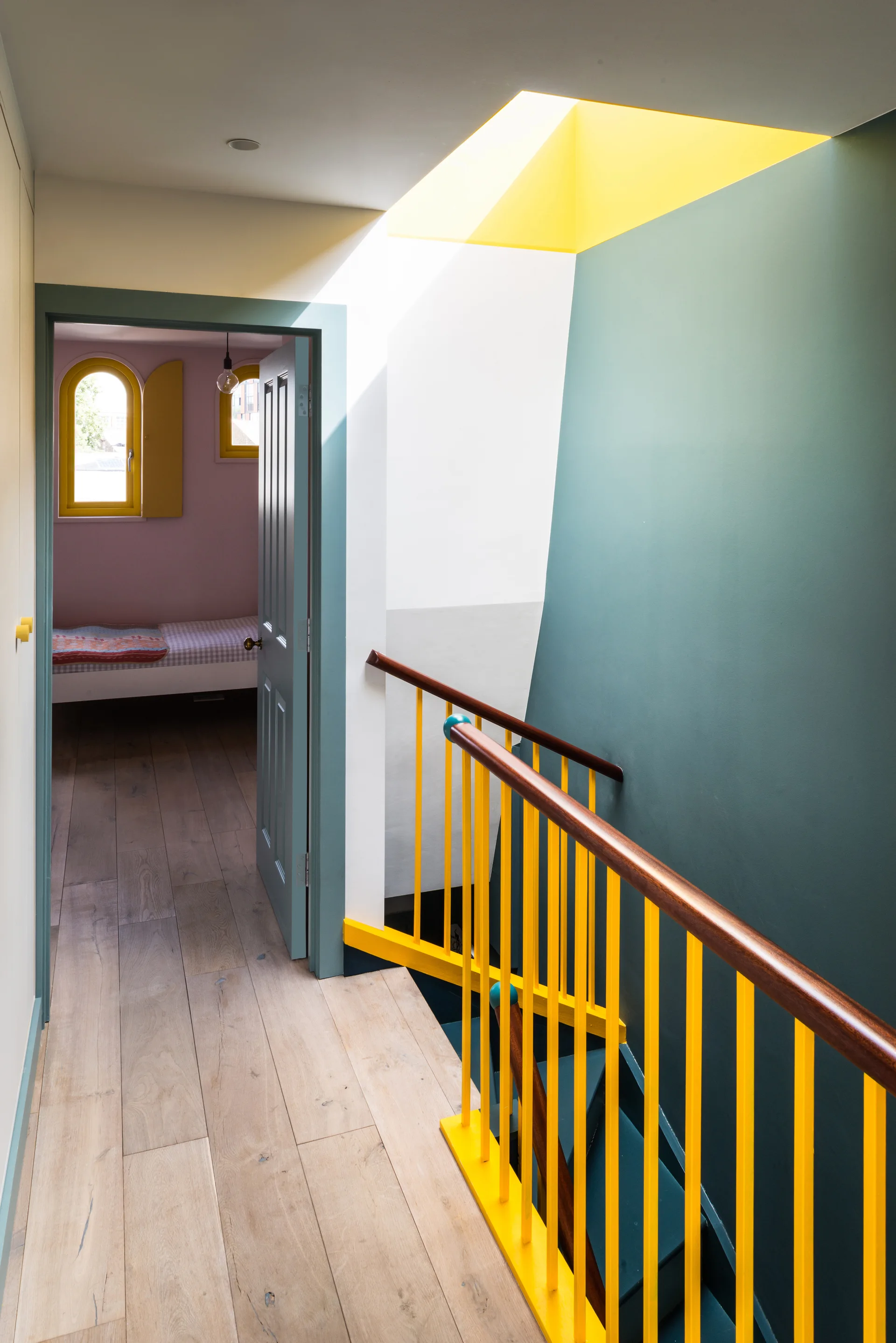 A modern residential hallway features teal walls, light wood flooring, a bright yellow staircase railing, and a view into a bedroom.