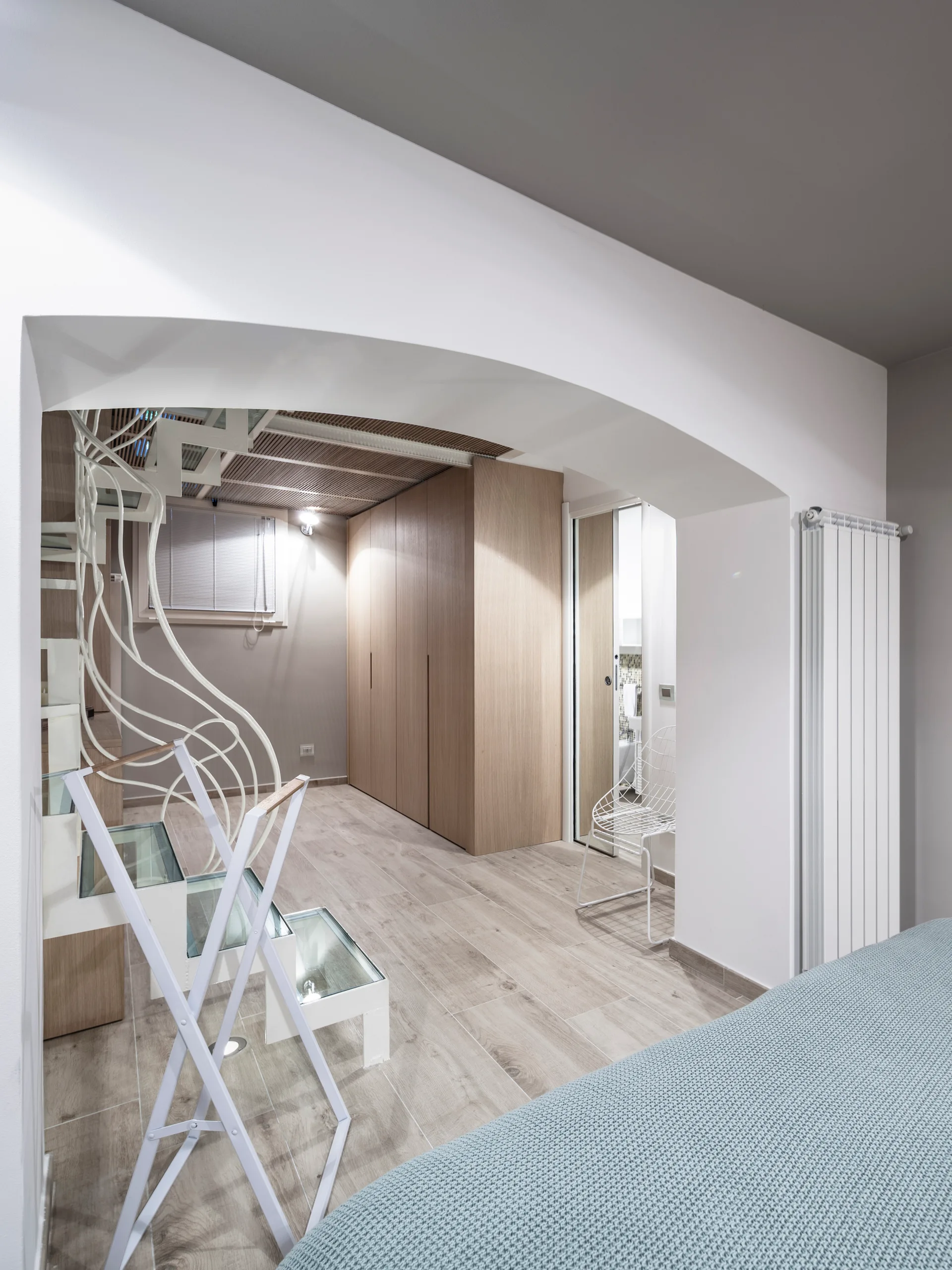 A bedroom view through a wide archway featuring a white metal staircase with glass treads and a seamless light wood wardrobe.