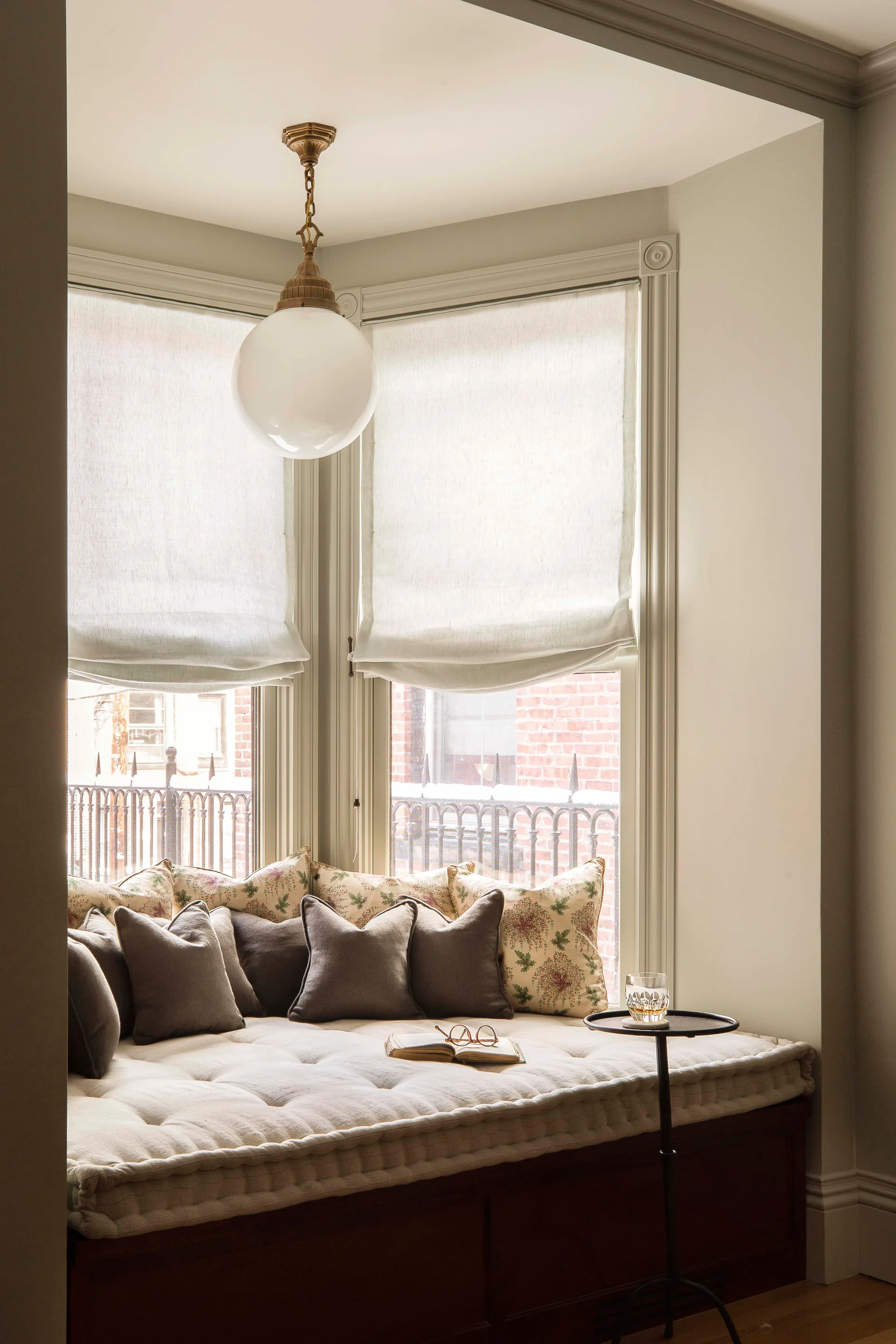 A cozy bay window seat features a tufted cream cushion, floral and solid taupe pillows, semi-sheer roman shades, and a brass globe pendant light.
