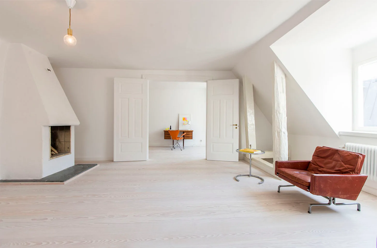 A bright minimalist living room featuring bleached wood floors, a plaster fireplace, a leather armchair, and open double doors.