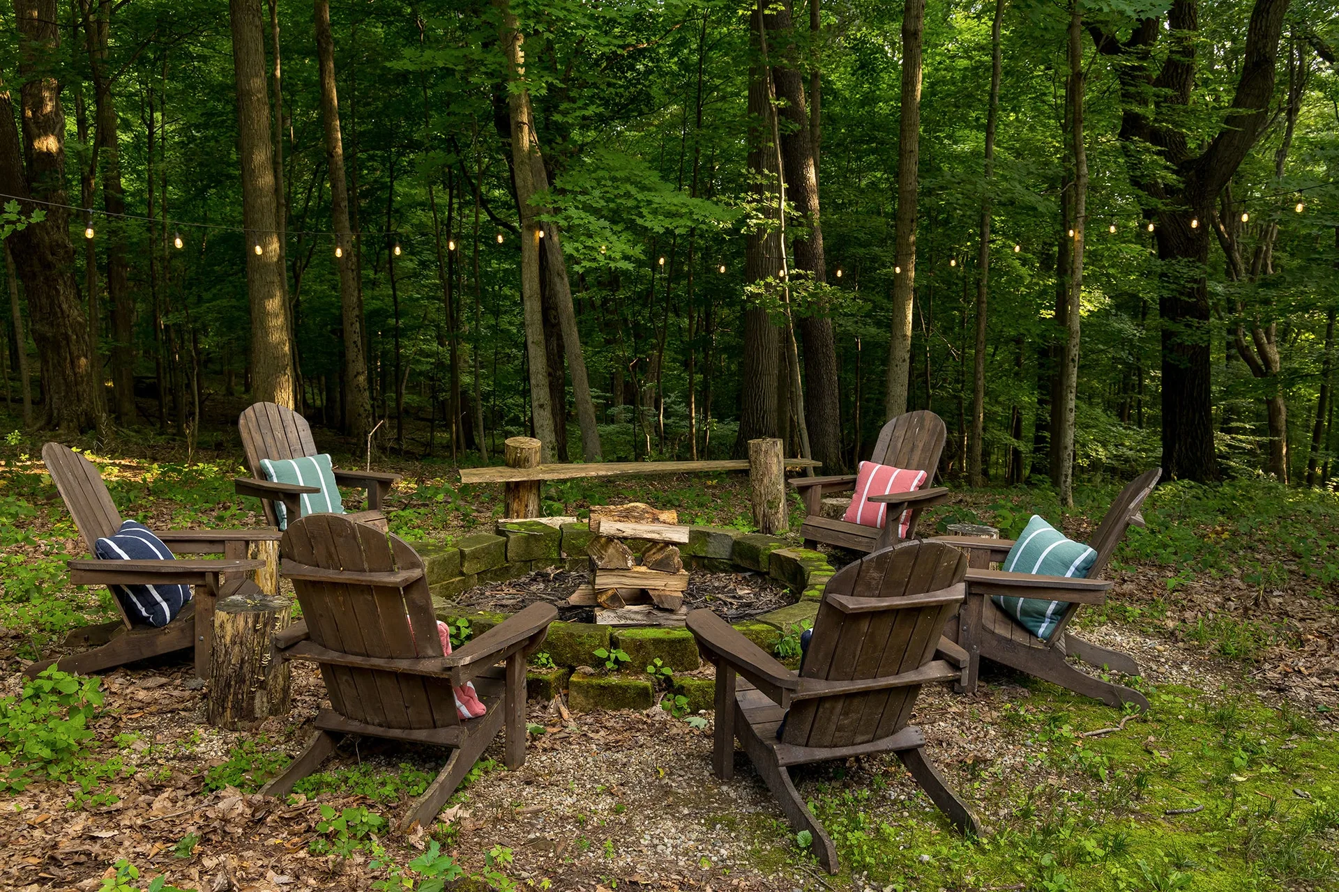 Dark-stained wooden Adirondack chairs with striped pillows surround a circular stone fire pit in a dense forest clearing under string lights.