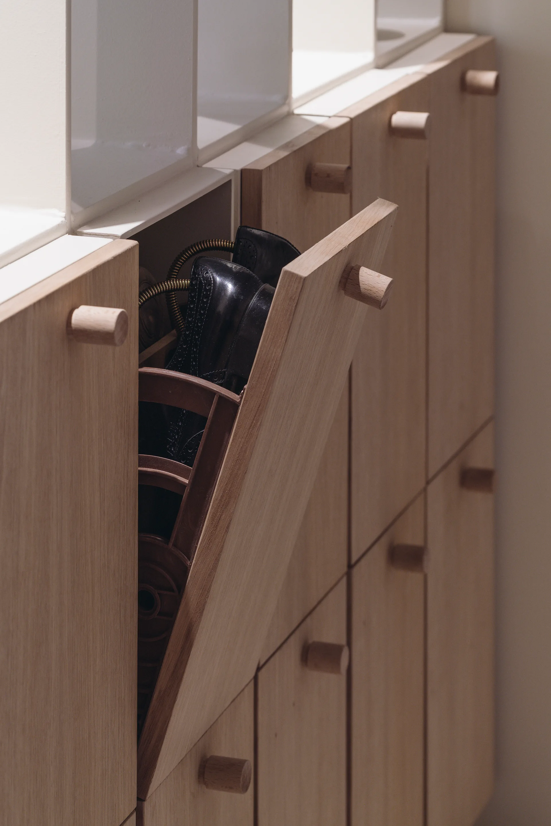 A close-up of a light wood entryway cabinet with a tilt-out drawer open, revealing a black shoe stored inside.