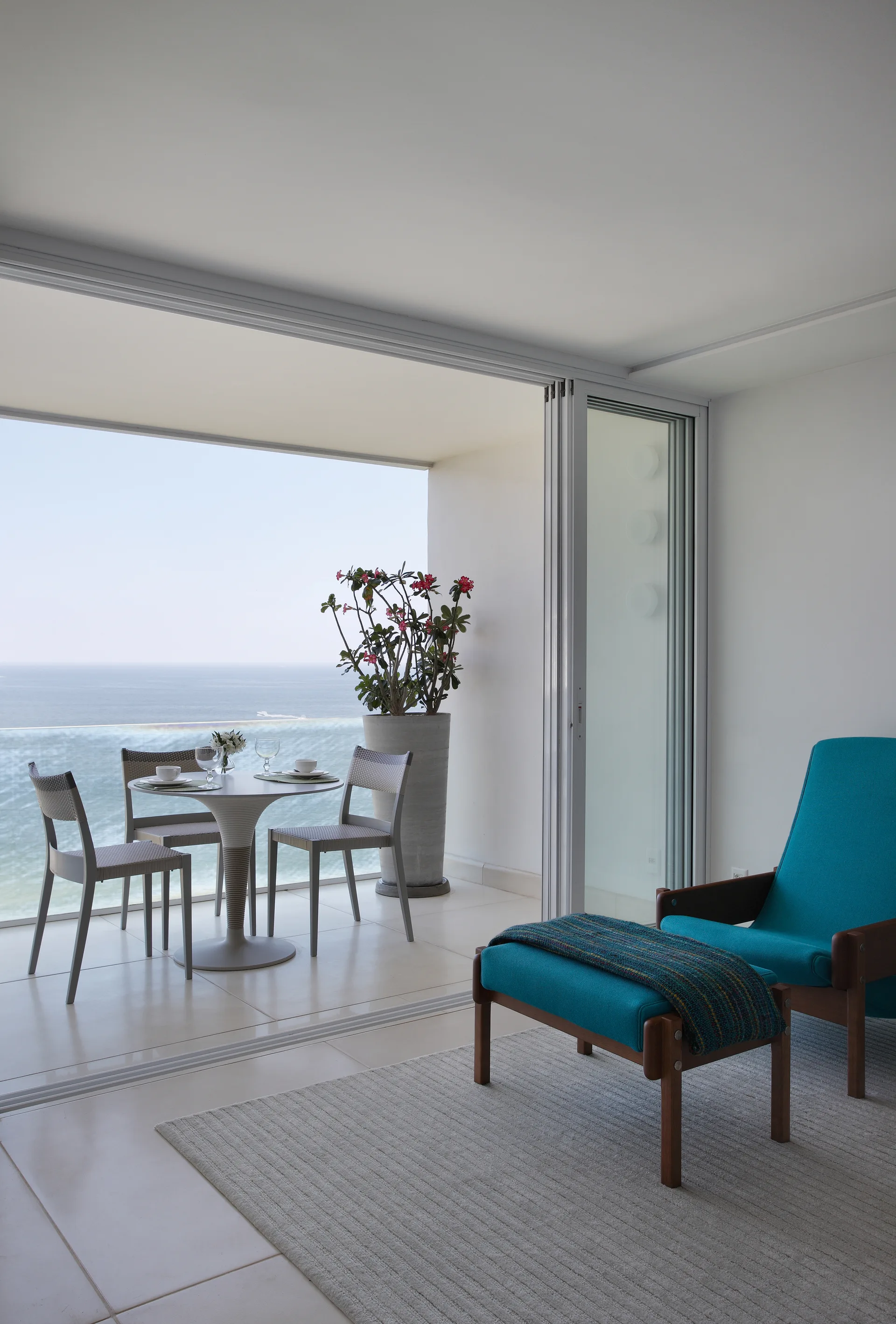 A teal lounge chair with an ottoman sits near sliding glass doors that open to a balcony dining set overlooking the ocean.