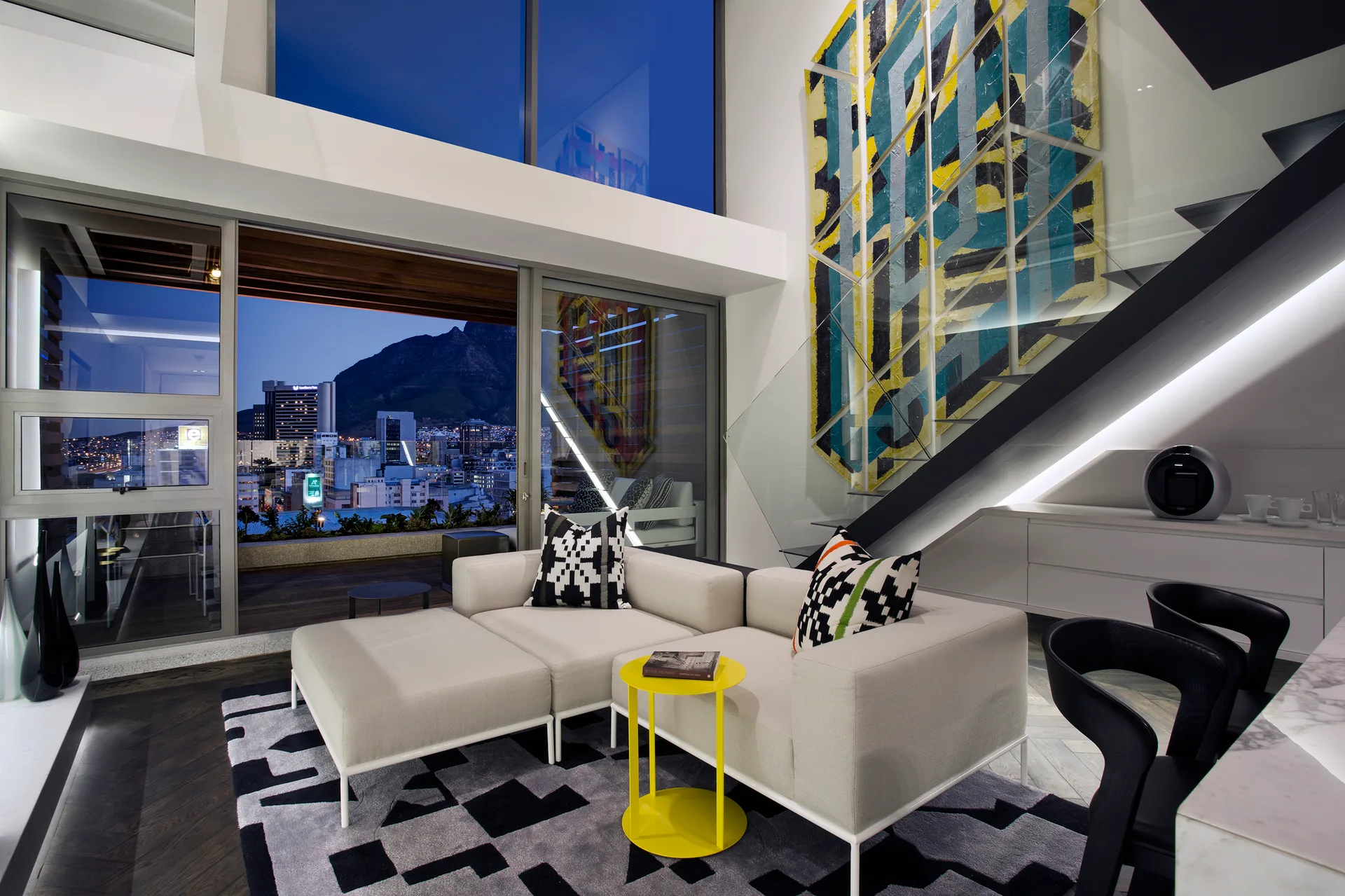 A modern living room features a light gray modular sofa on a geometric rug next to a floating staircase with glass railing.