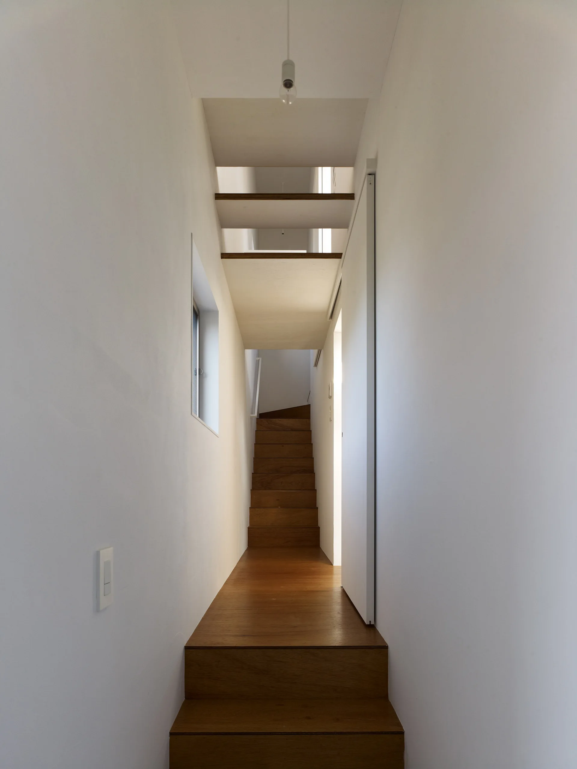 A minimalist narrow hallway featuring a straight wooden staircase, white walls, and open wood joists across a recessed ceiling.