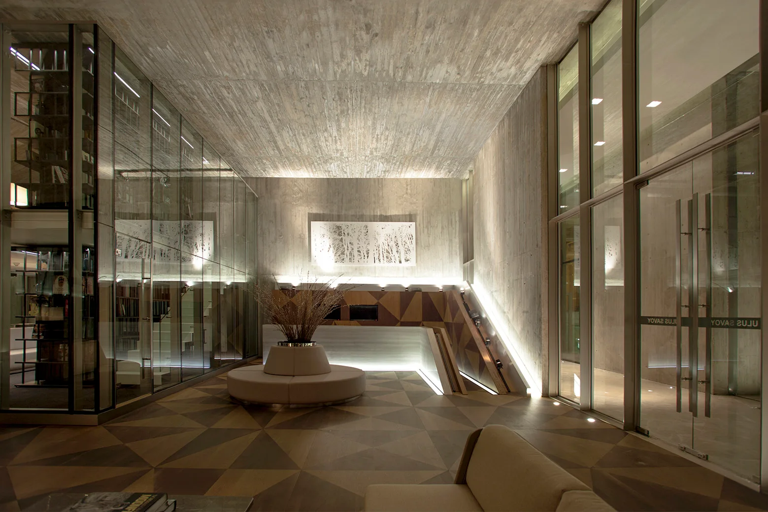A modern lobby with board-formed concrete walls and ceiling features a circular bench, a wood-paneled reception desk, and glass partition walls.