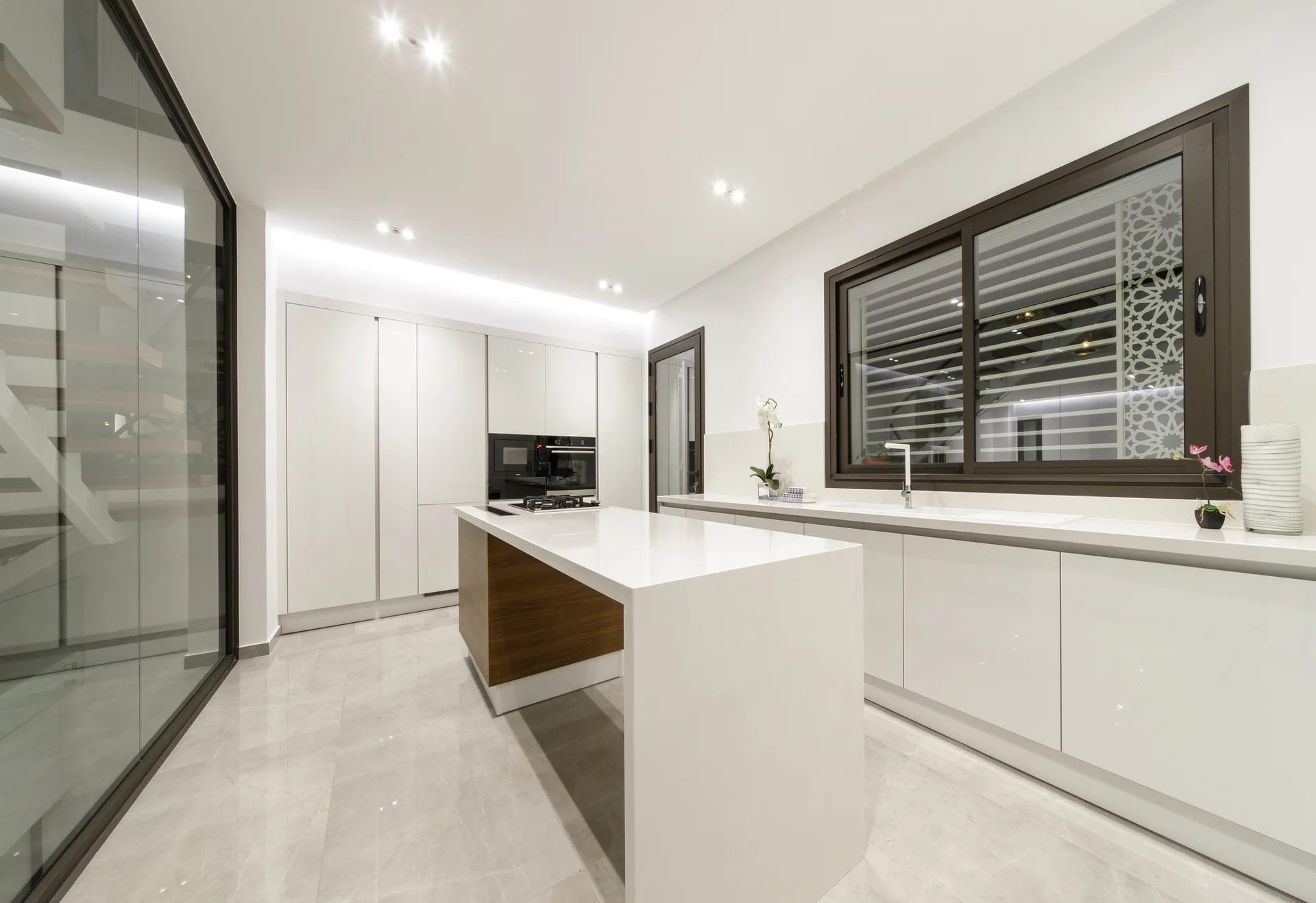 A minimalist white kitchen features a central waterfall island with a wood base, glossy cabinetry, and polished grey tile flooring.