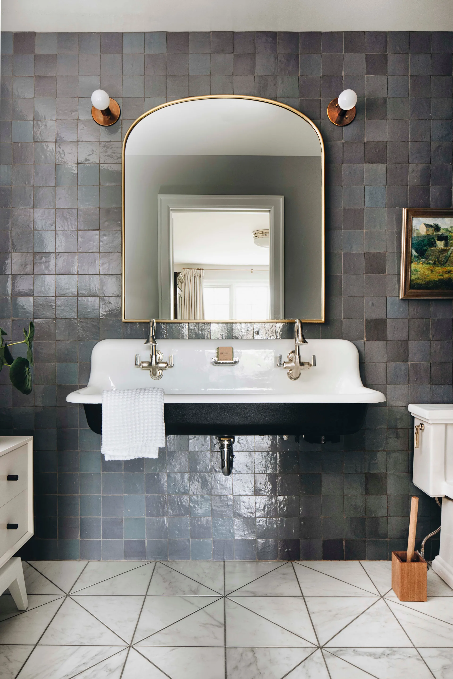 A bathroom featuring a wall-mounted white sink with black trim set against a dark grey square tiled wall beneath an arched brass mirror.