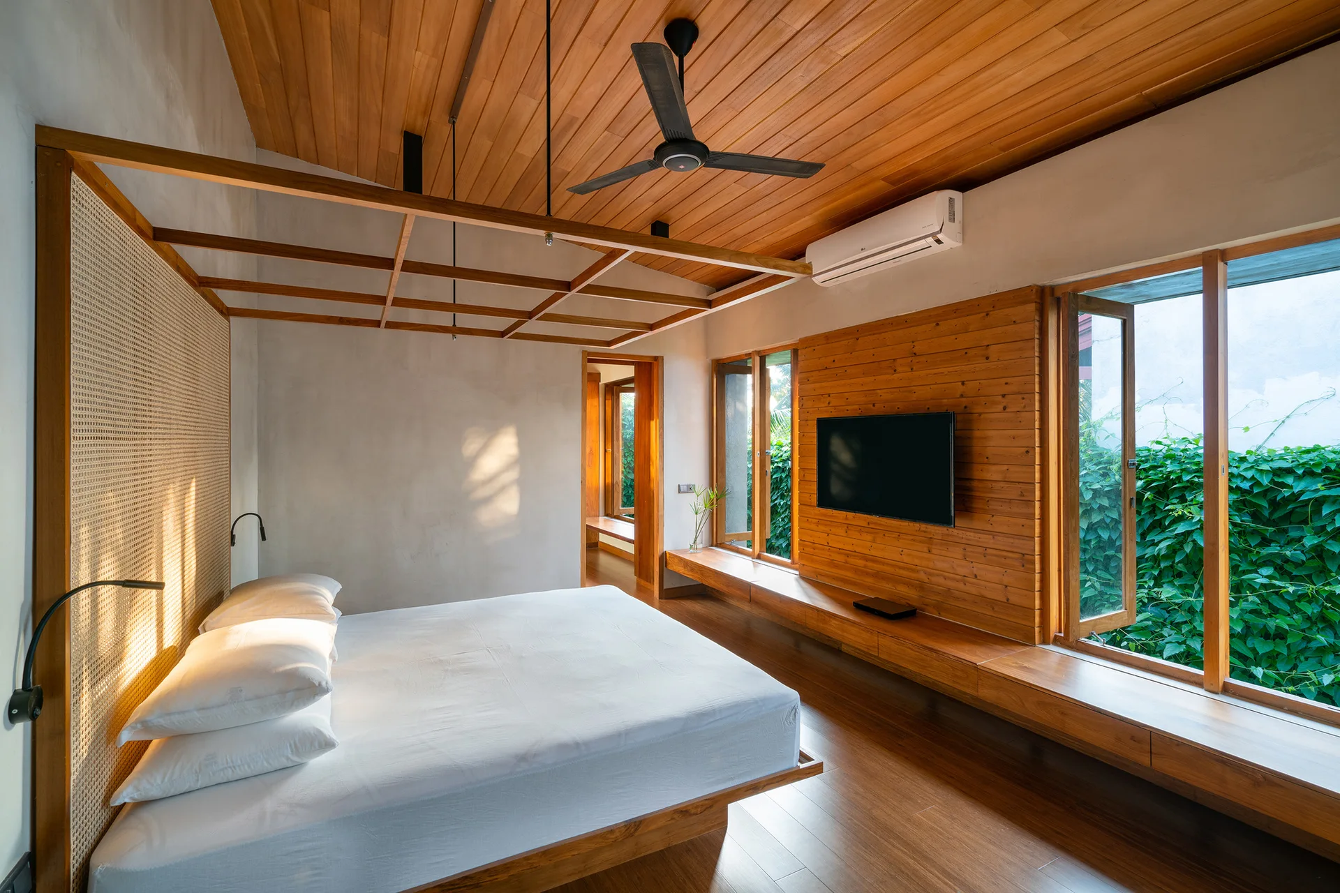 A modern bedroom features a low bed with a wooden canopy frame, a wood plank ceiling, and a wall-mounted TV on a paneled accent wall.