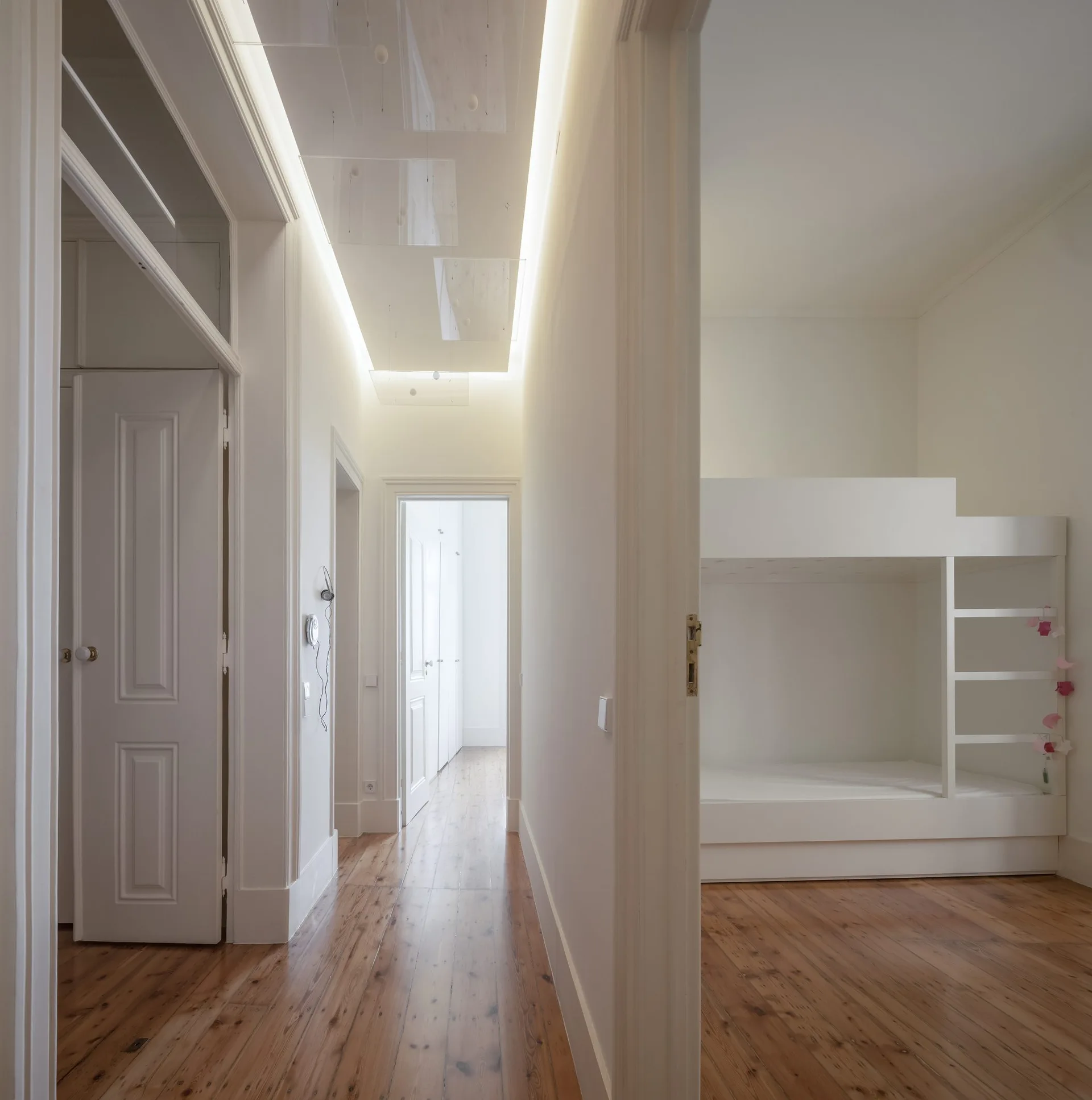 A white hallway with polished wood flooring and a recessed ceiling cove light leads past a bedroom containing a minimalist bunk bed.