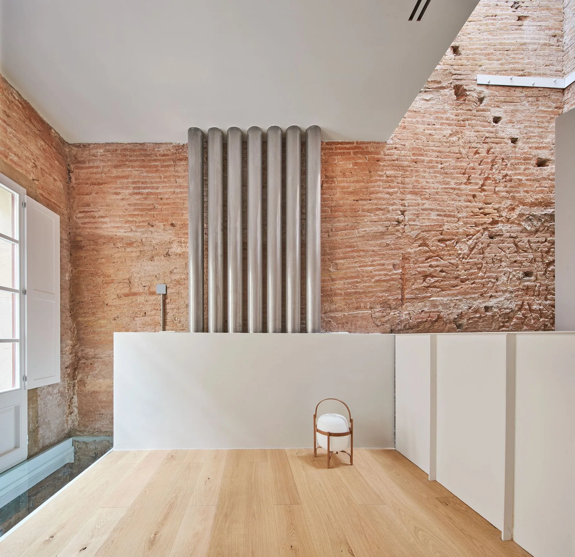 A minimalist room features an exposed red brick wall, vertical metallic pipes, a light wood floor, and sleek white low-profile cabinetry.