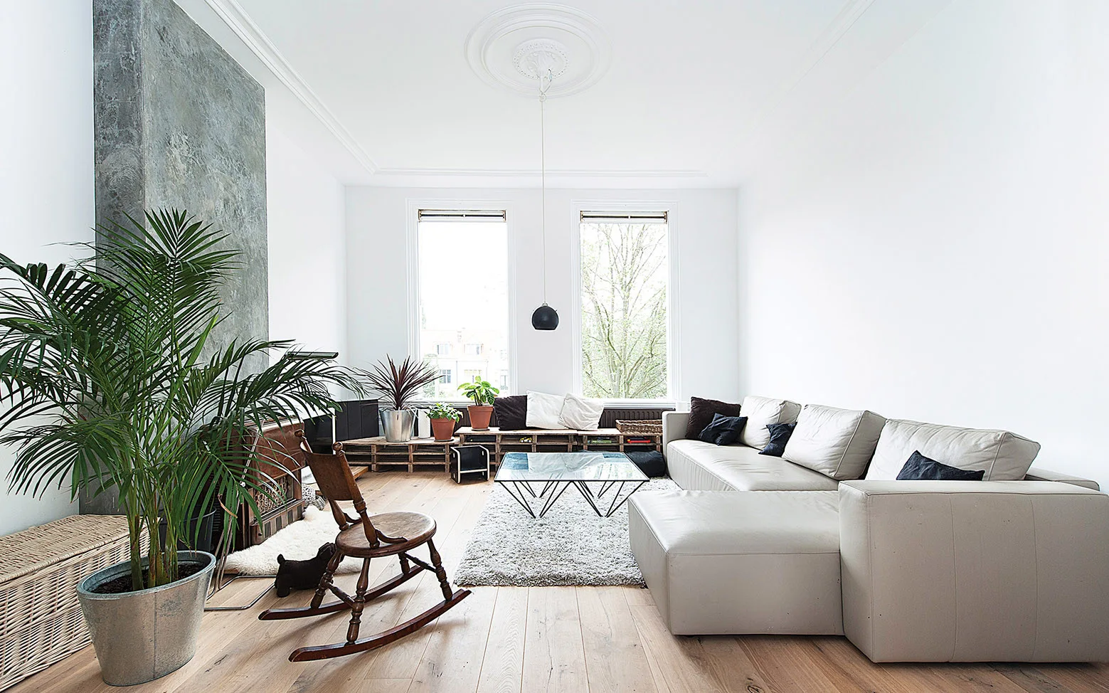 A bright white living room with light wood floors, a large grey sectional sofa, an antique rocking chair, and a tall textured grey fireplace.